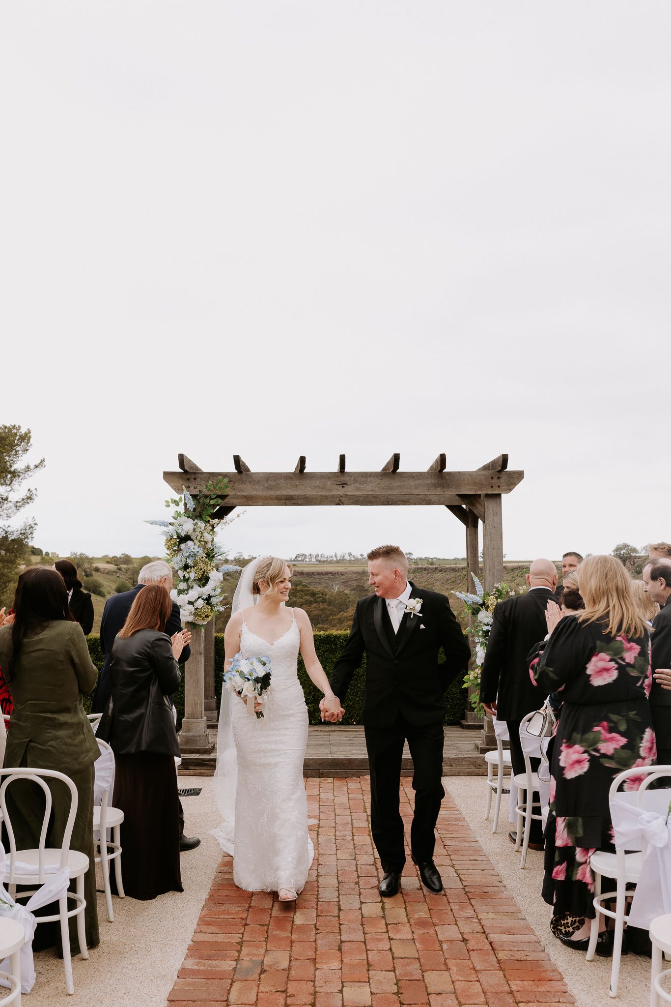 Wedding guests stand during an outdoor ceremony on a brick patio under a wooden pergola.