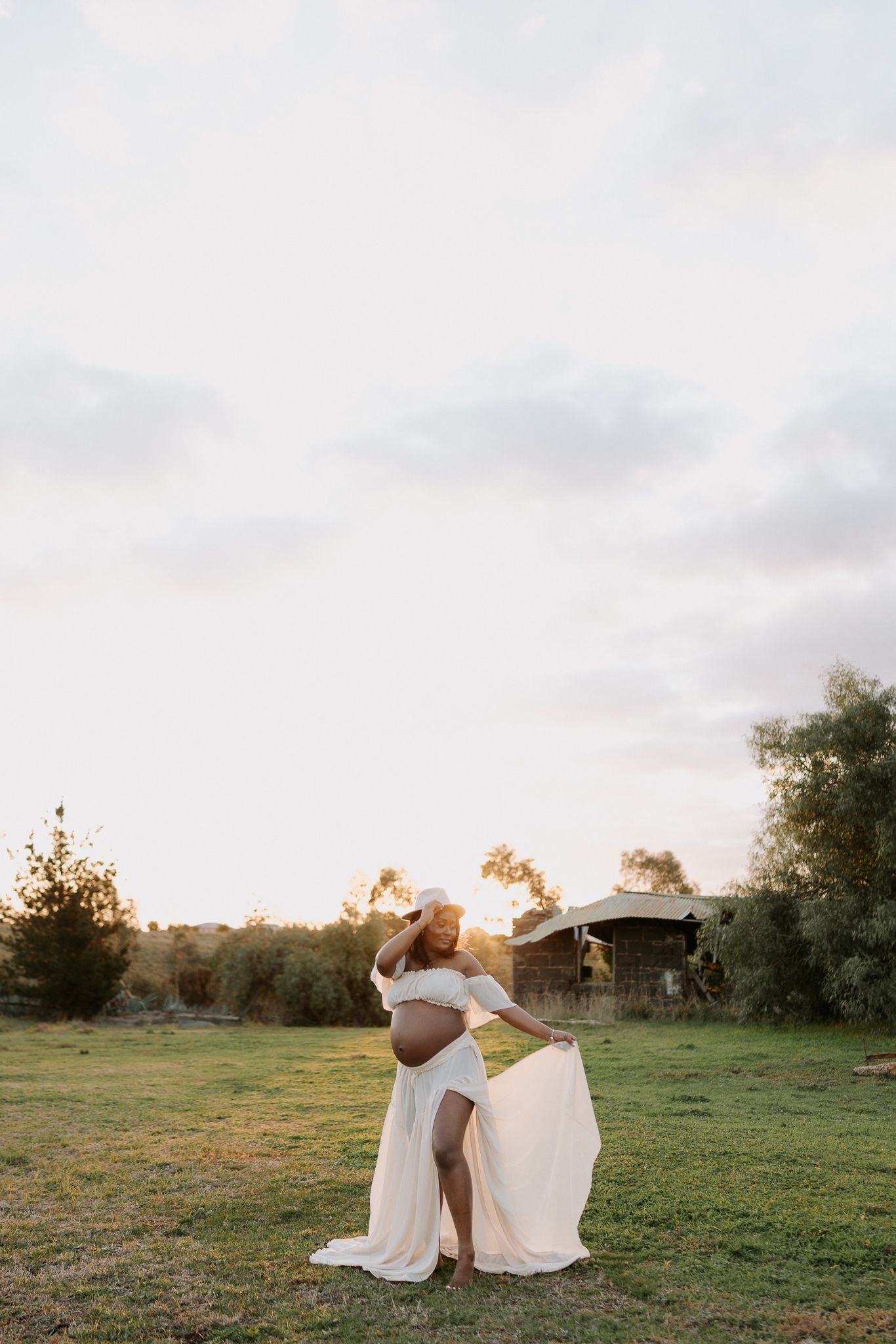 A pregnant woman in white flowing dress stands in a grassy field at sunset with golden light behind her.