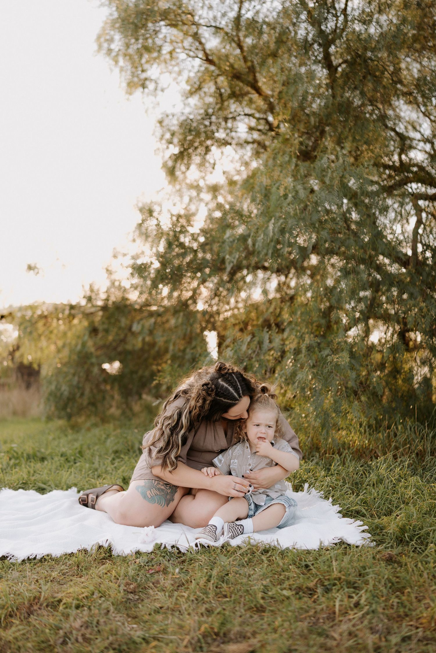 Mother sitting on white blanket outdoors, lovingly embracing her baby near lush green trees in golden hour light.