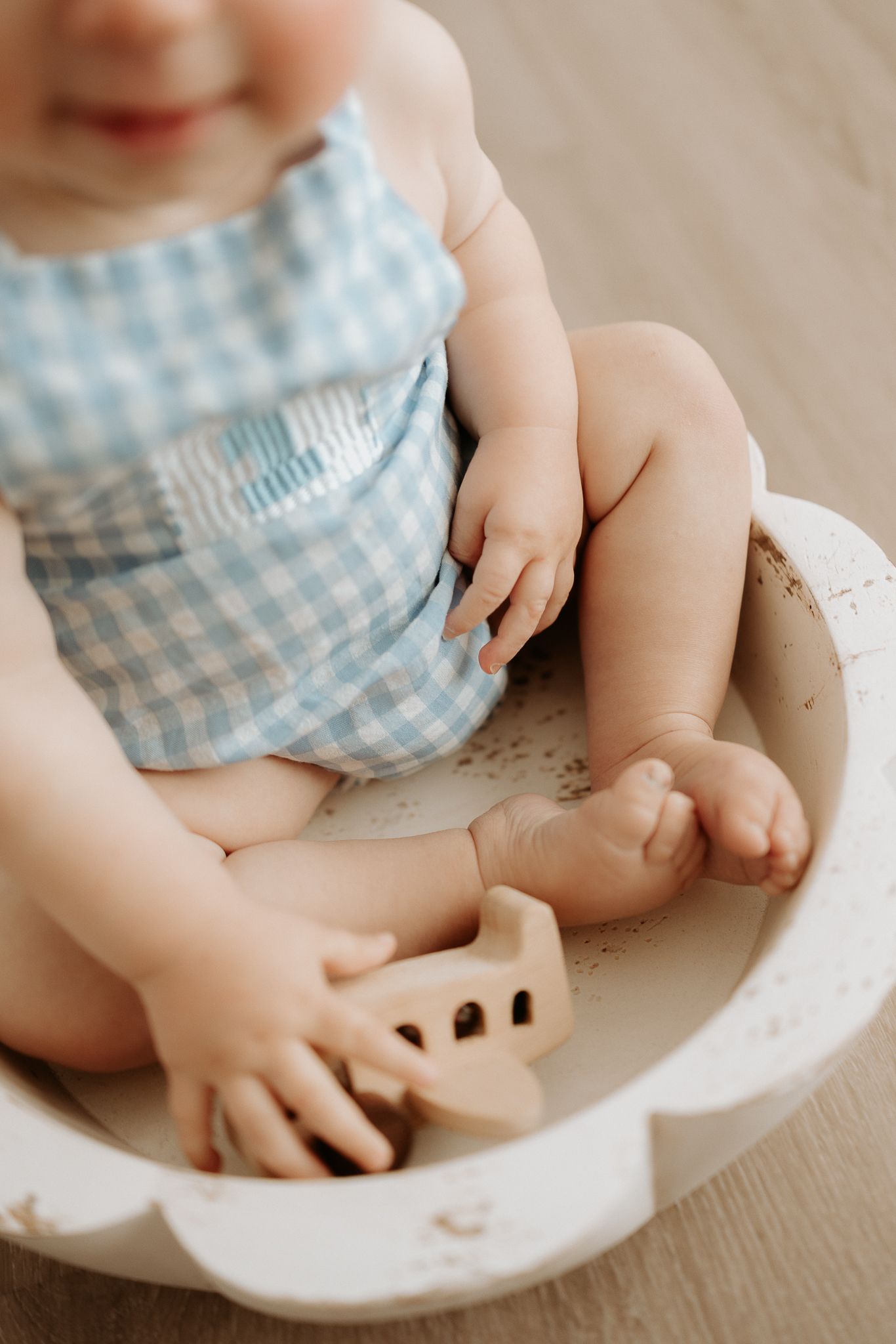 Baby in blue gingham romper sitting in a white wooden bowl, holding a wooden toy airplane.