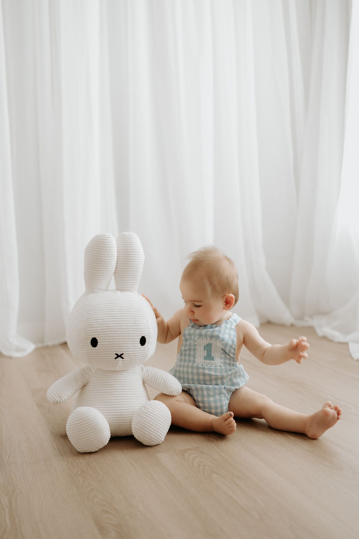 Baby in blue gingham romper sitting on wood floor next to large white Miffy bunny plush toy.