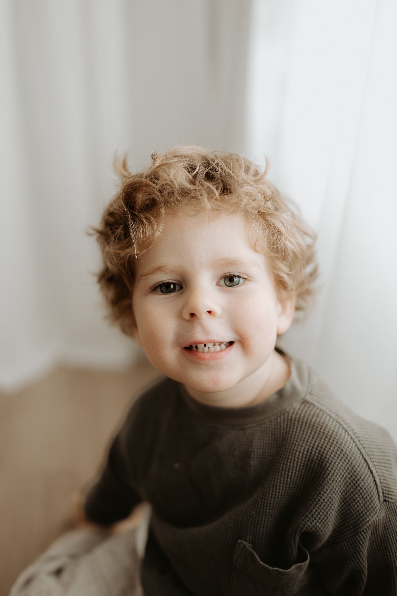 Adorable toddler boy with curly blonde hair smiling cheerfully, wearing a dark olive green top.