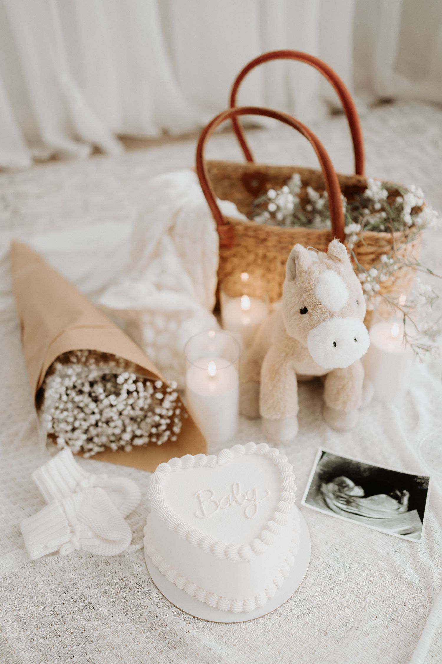 White baby shower display with heart cake, unicorn toy, ultrasound photo, candles, flowers, and wicker basket on white fabric.