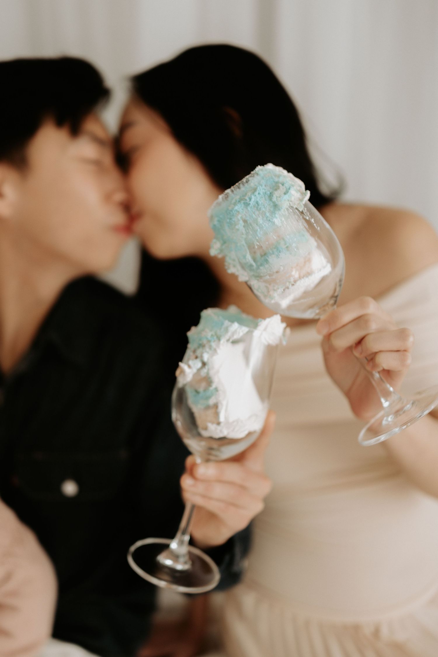 Couple kissing while holding wine glasses decorated with blue and white cotton candy in a romantic setting.