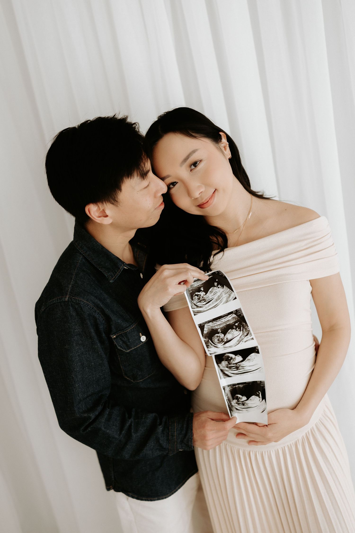 Expecting Asian couple holding ultrasound photos, woman in off-shoulder dress, man in denim shirt, maternity announcement portrait.