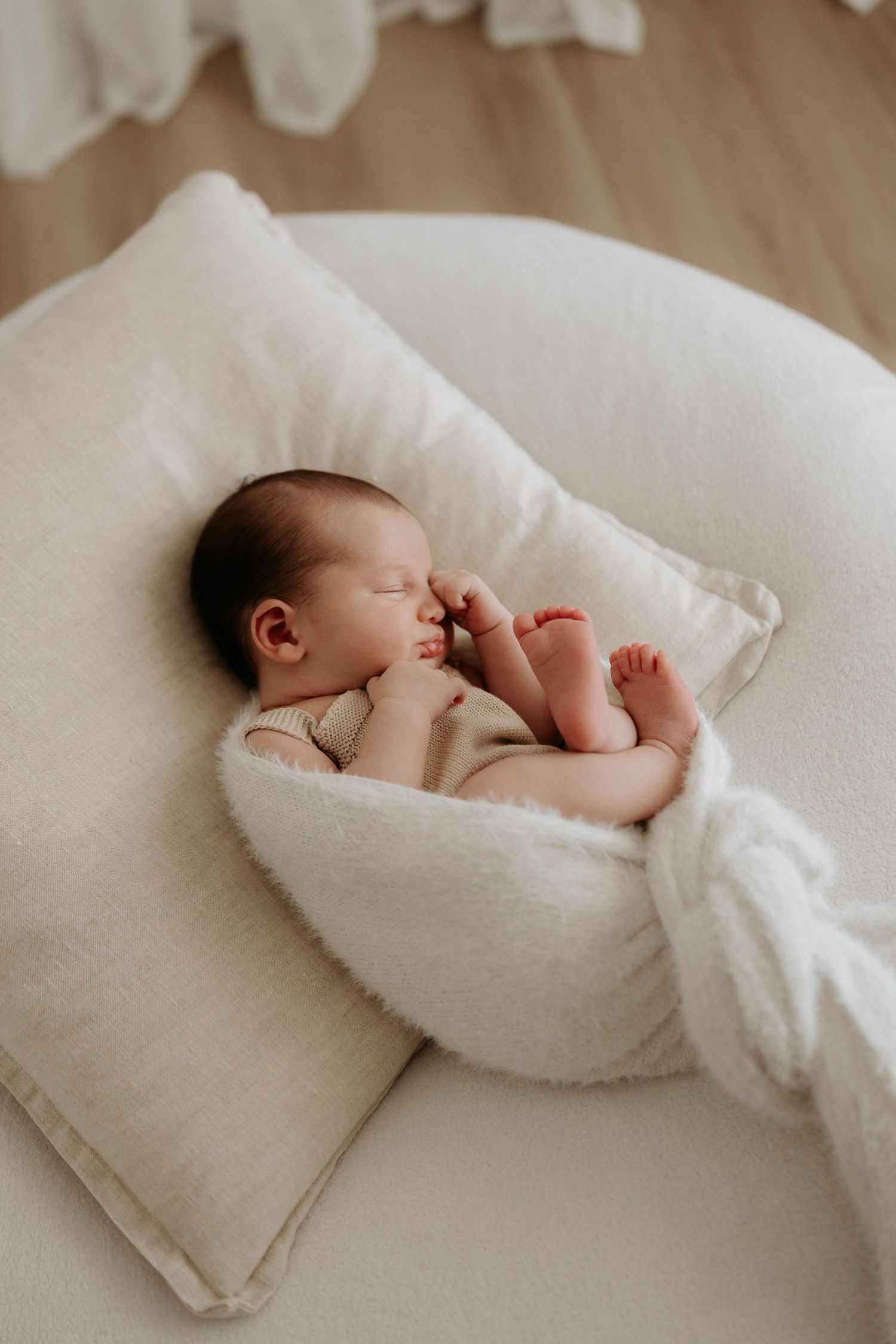 Newborn baby sleeping peacefully in a white fluffy nest pillow, wrapped in a soft cream blanket.