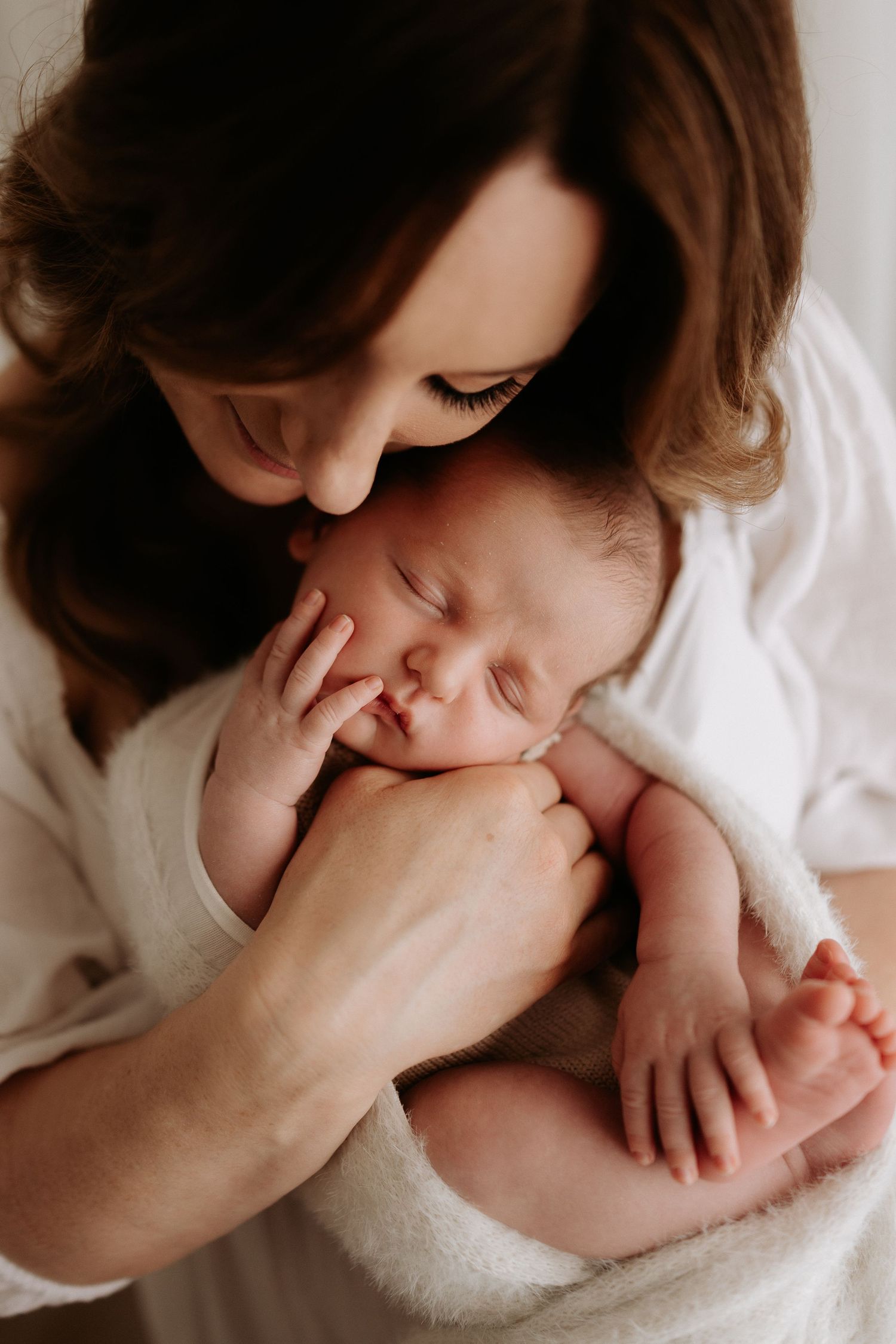 A mother tenderly kisses her sleeping newborn baby wrapped in a soft white blanket.