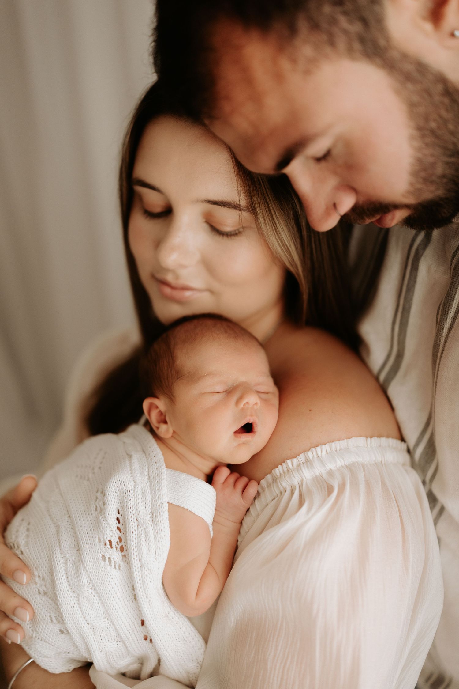 New parents tenderly hold their sleeping newborn baby wrapped in a white knit blanket in a soft, natural light setting.