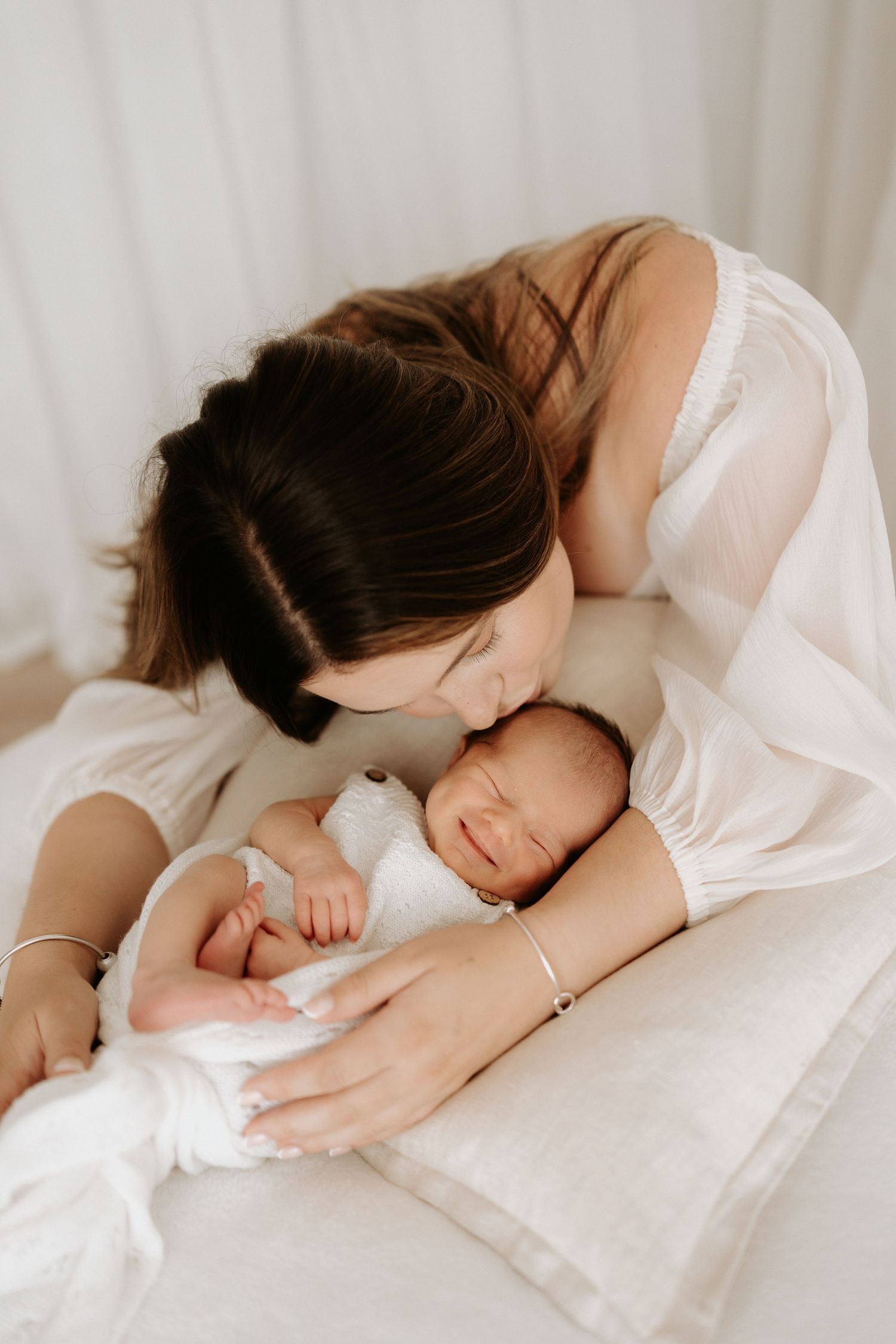 A tender mother in white dress gently kisses her sleeping newborn baby wrapped in white cloth on a soft bed.
