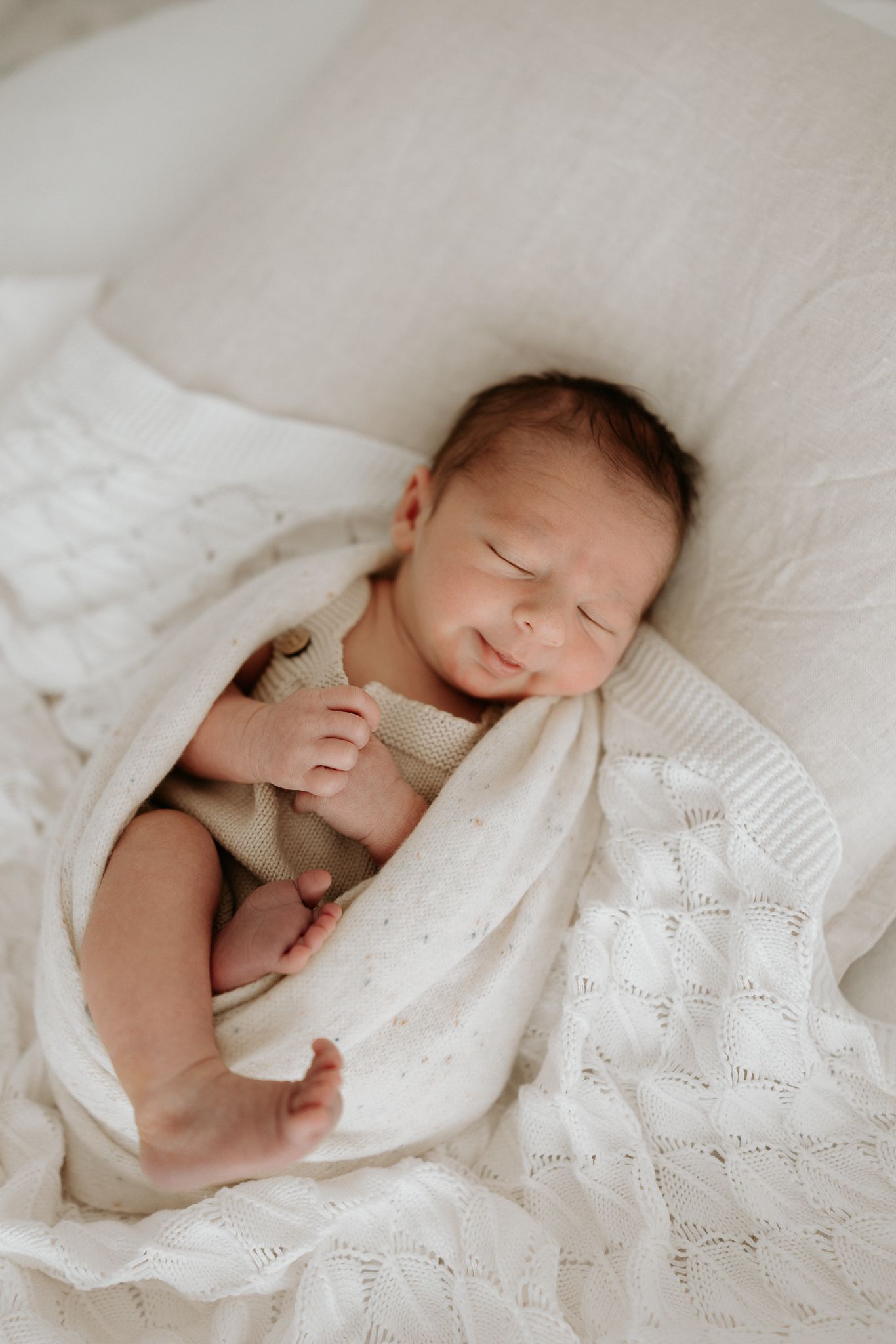 Peaceful newborn baby sleeping wrapped in a soft white knit blanket, wearing a beige onesie on a white bed.