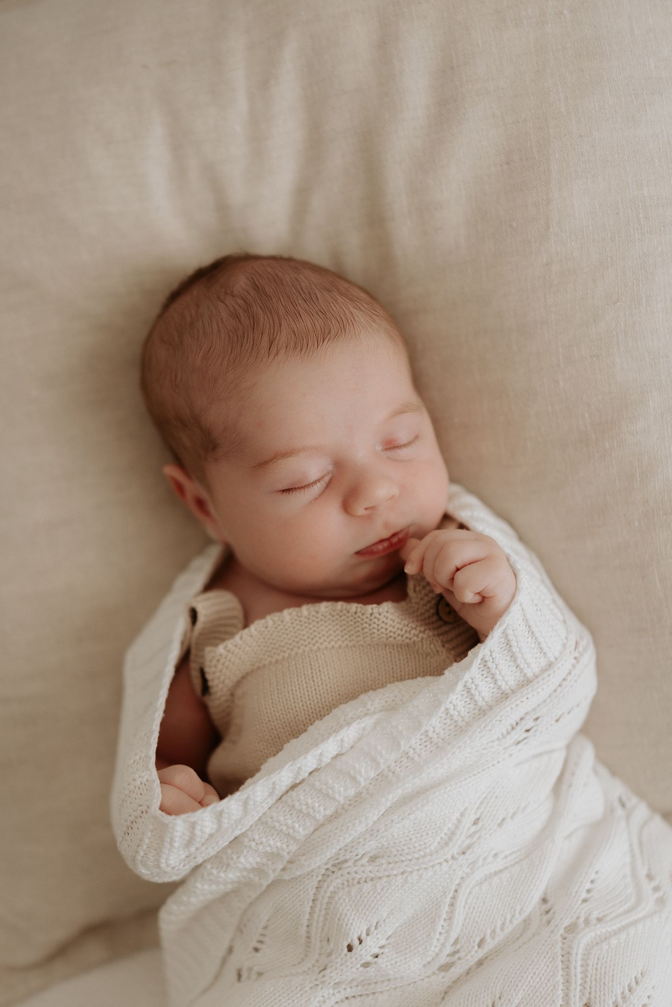 Peaceful newborn baby sleeping wrapped in a white cable-knit blanket on a soft beige background.