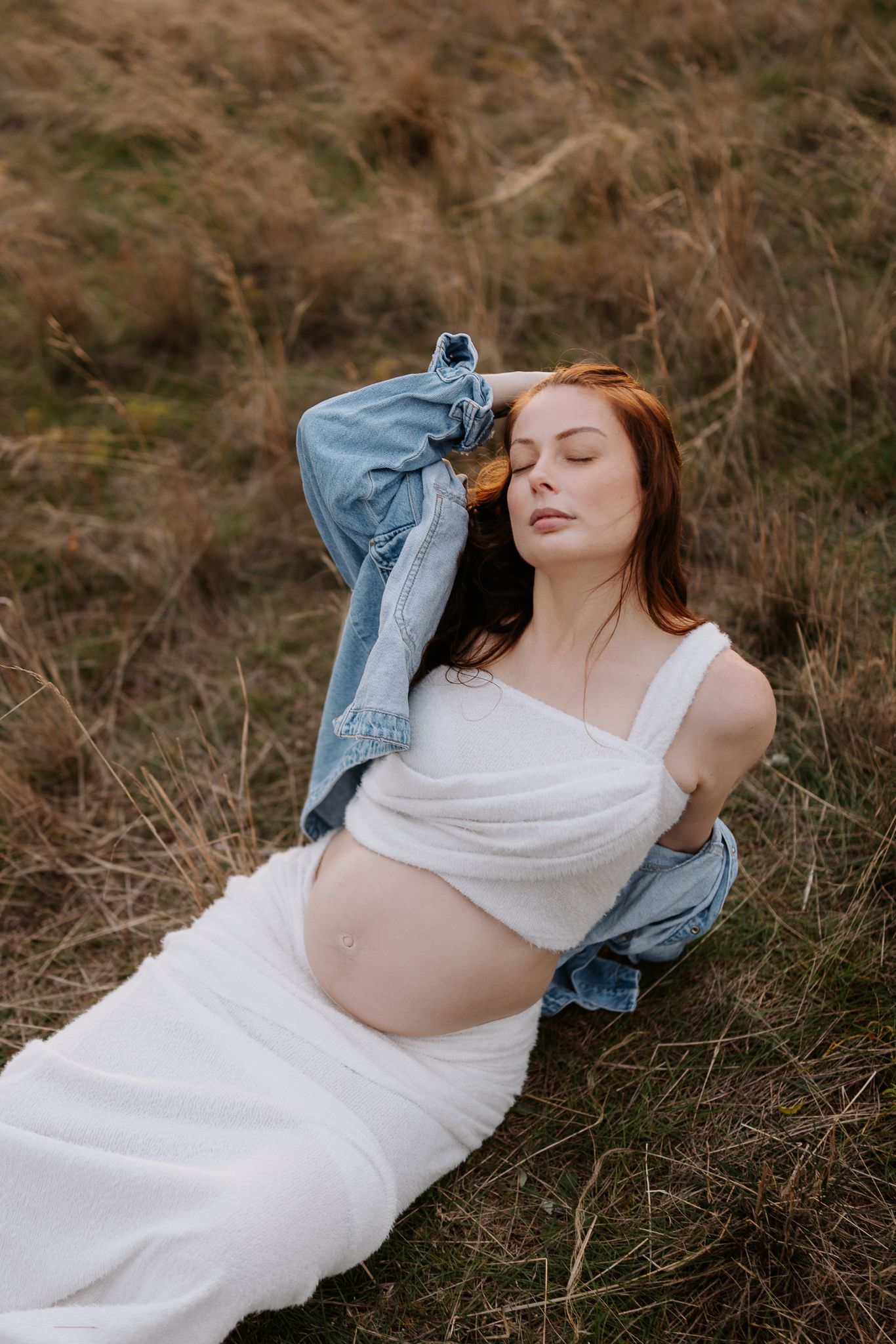 Pregnant woman in white dress and denim jacket resting in a field with dry grass, eyes closed, cradling her baby bump.