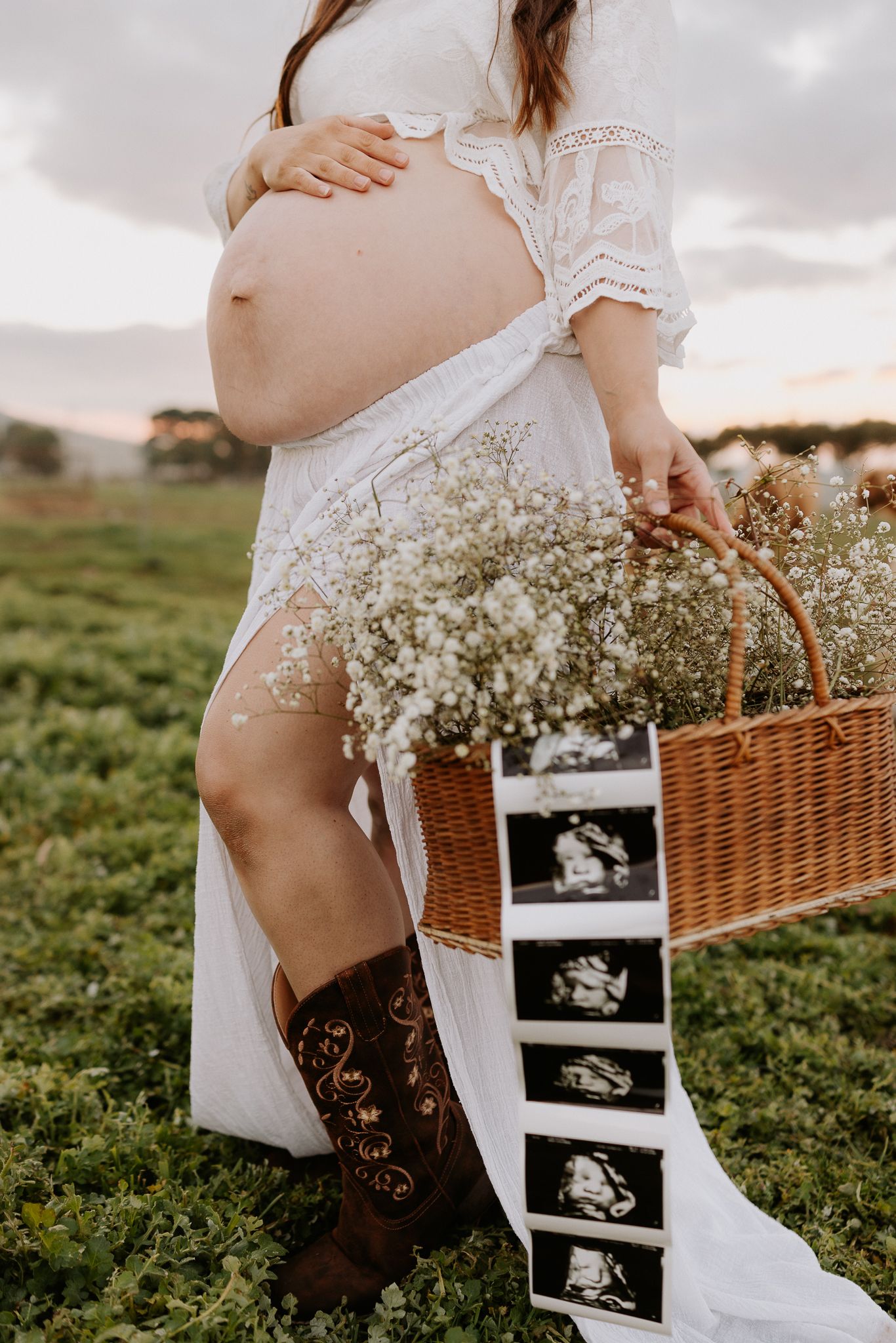 Pregnant woman in white lace dress and cowboy boots holding wicker basket with baby's breath flowers and ultrasound photos outdoors.