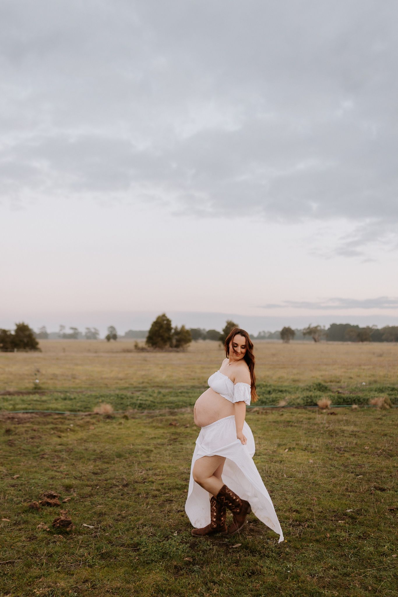 Pregnant woman in white off-shoulder dress posing in green field at dusk, showcasing baby bump.