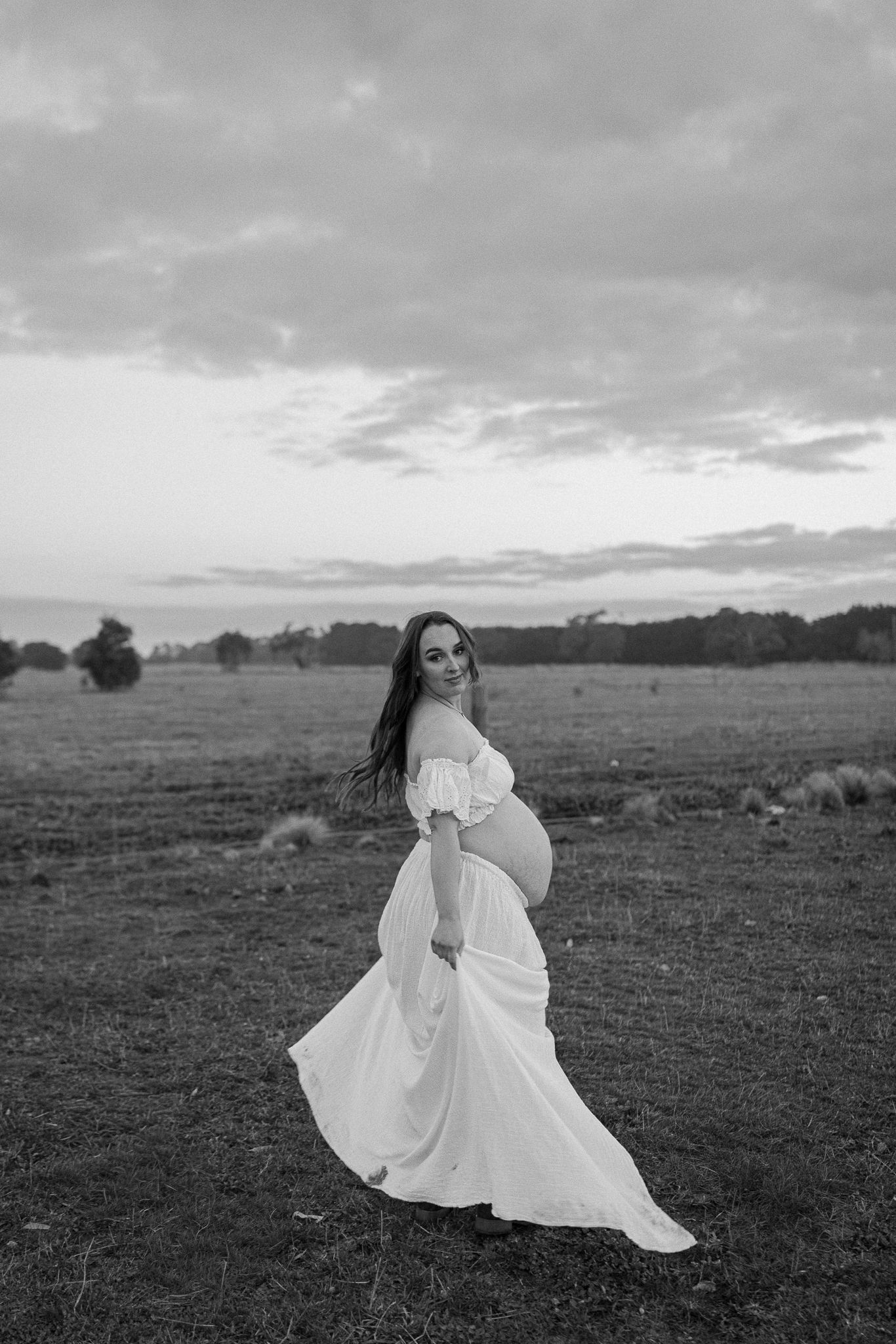 Black and white photo of a pregnant woman in a flowing white off-shoulder gown posing gracefully in an open field.