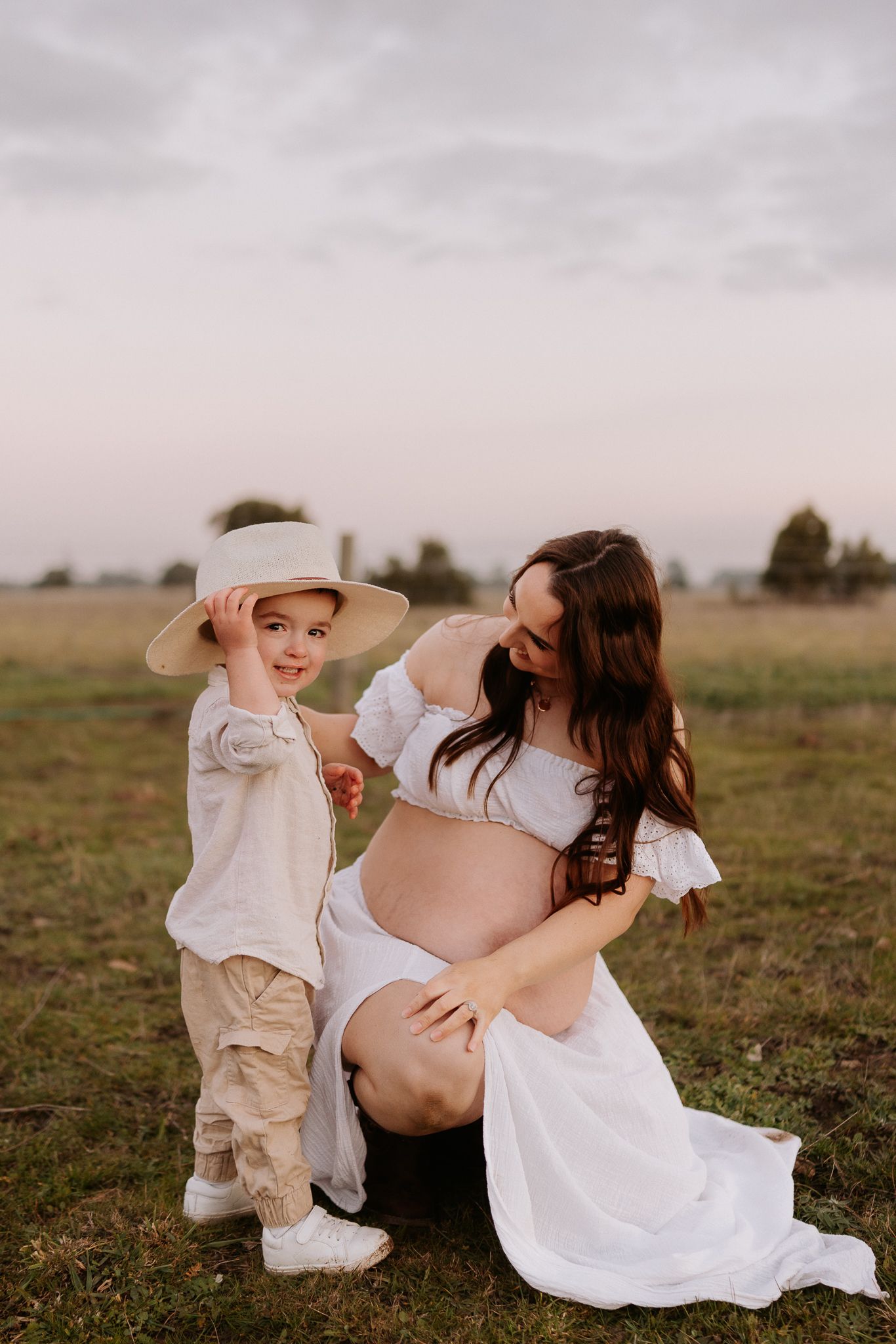 Pregnant woman in white dress kneeling beside young boy wearing hat in open field at sunset.