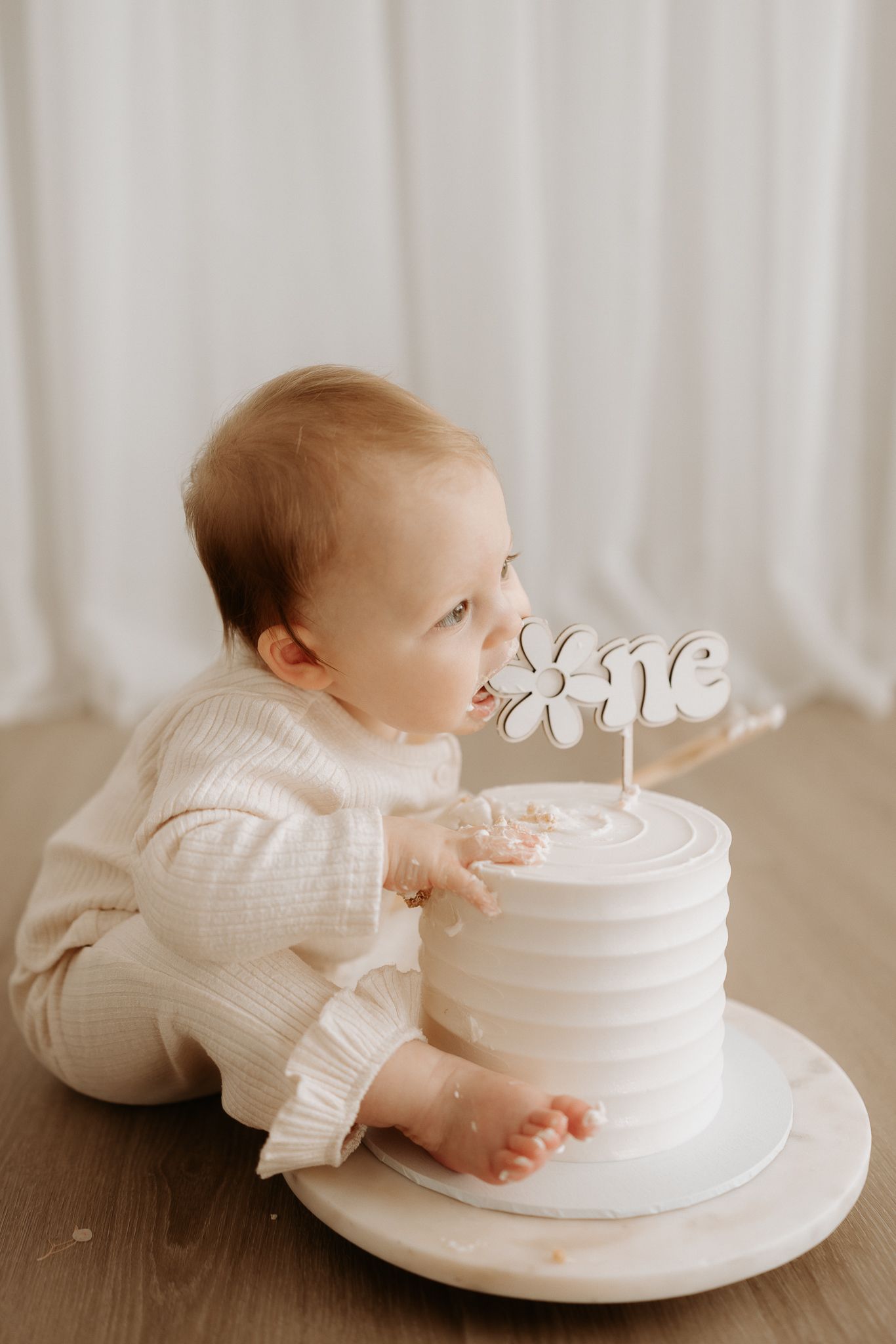Baby in cream outfit smashing a white first birthday cake with a floral 'one' topper on a wooden board.