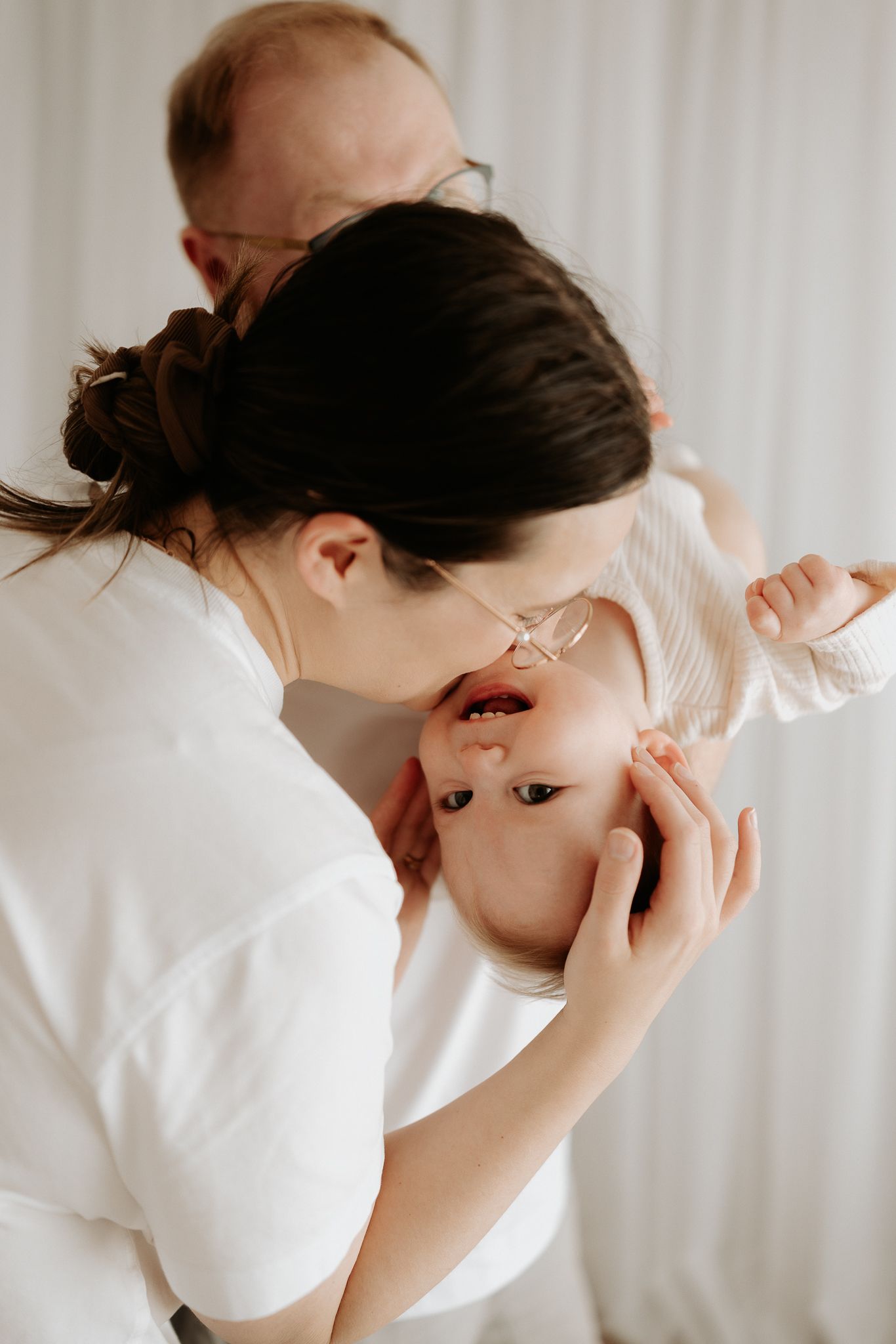 A mother kisses her baby while a father looks on tenderly in a bright, minimalist white setting.