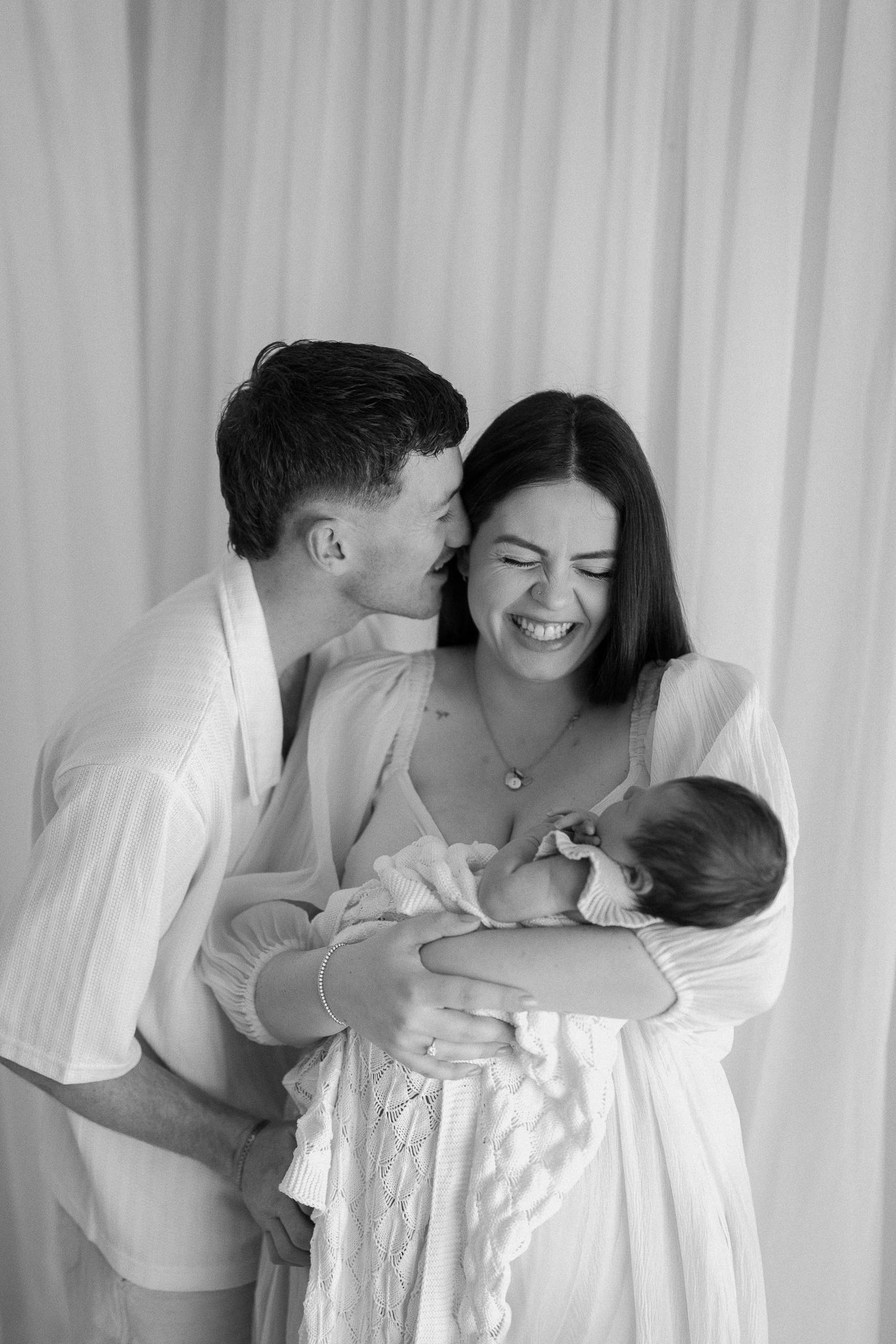 Black and white photo of happy couple with newborn baby, man kissing woman's cheek as she laughs.