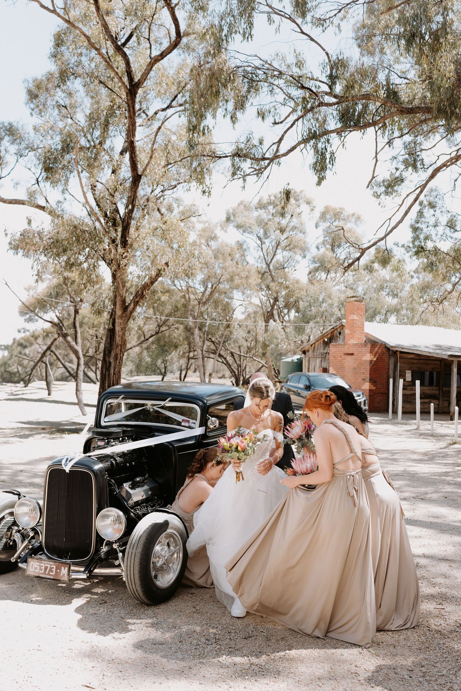 A classic black vintage car parked with people in formal attire and flowing gowns gathered around it in a rustic outdoor setting.