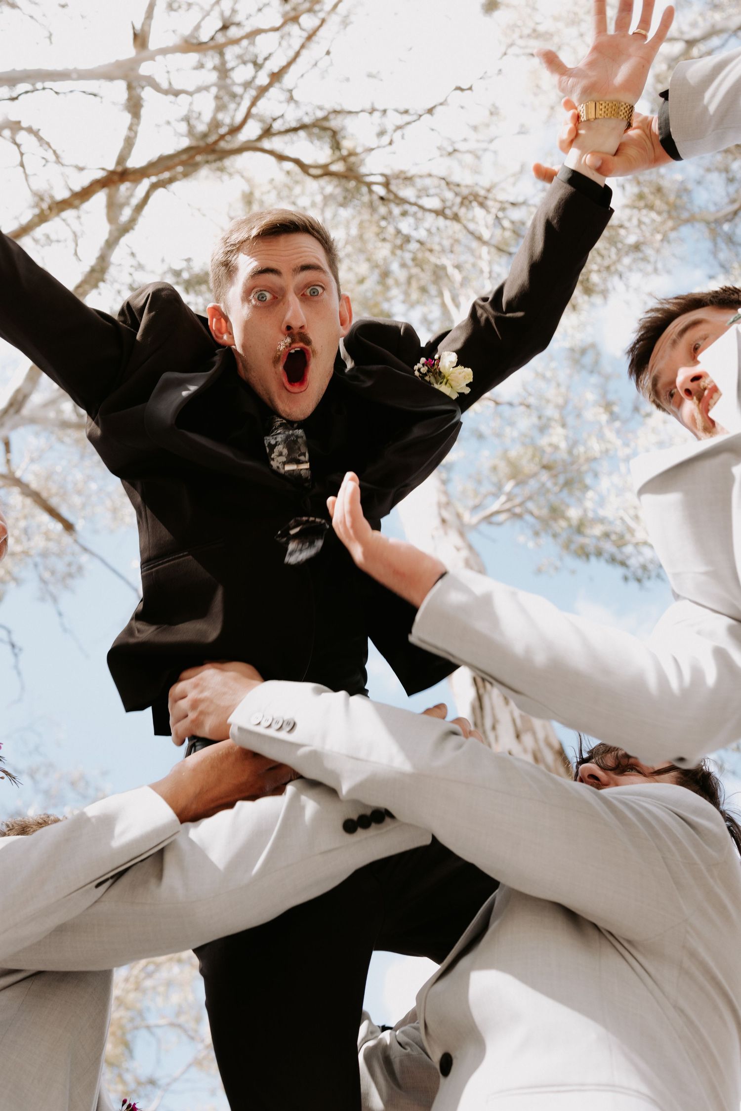 Energetic groomsmen in black and white suits jumping and celebrating outdoors with tree branches in background.