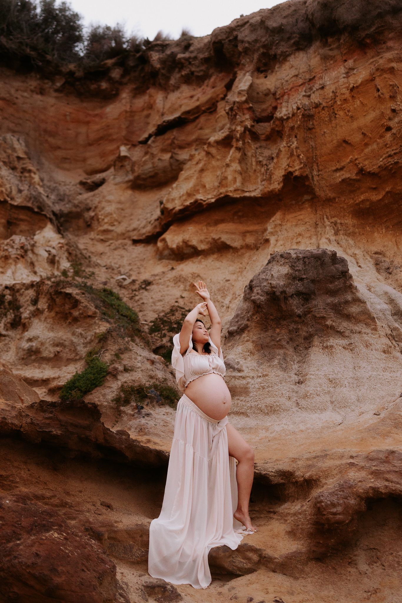 Pregnant woman in white flowing dress poses gracefully against rustic sandstone cliffs outdoors.