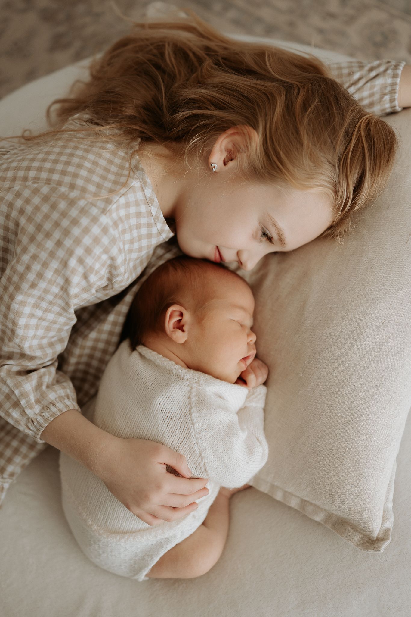 A young mother with wavy blonde hair tenderly rests her cheek against her sleeping newborn baby wrapped in white.