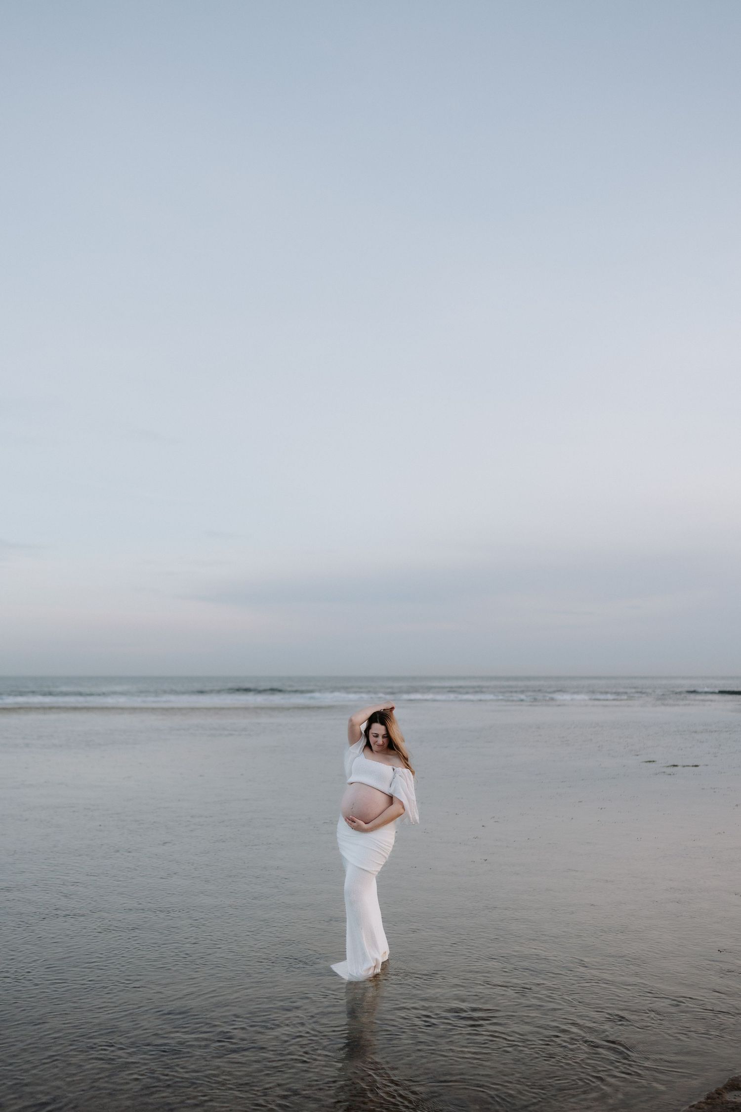 Pregnant woman in white off-shoulder gown standing in shallow ocean water at sunset during a maternity photoshoot.