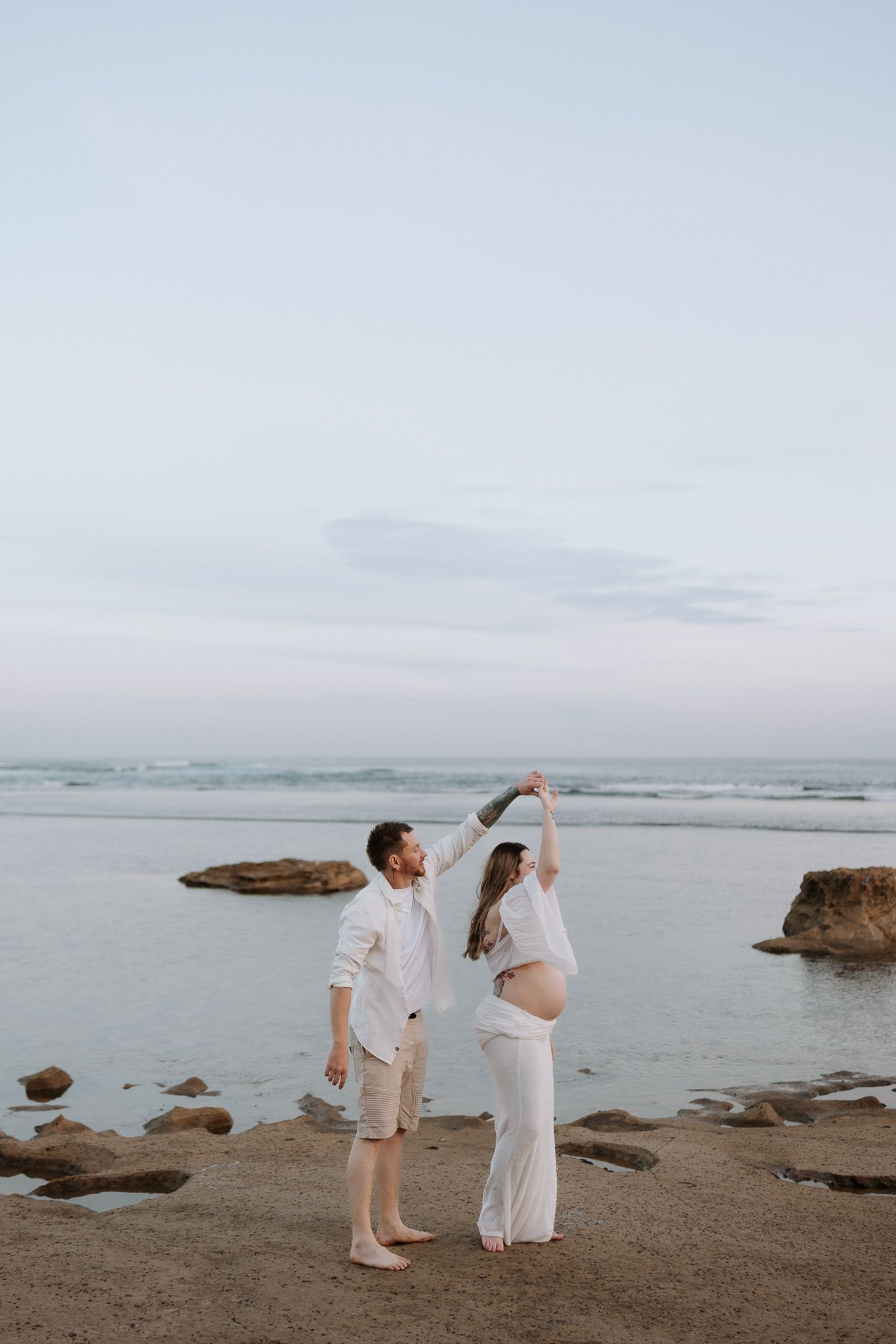 Couple dancing on beach at sunset, pregnant woman in white outfit held by partner near ocean rocks.