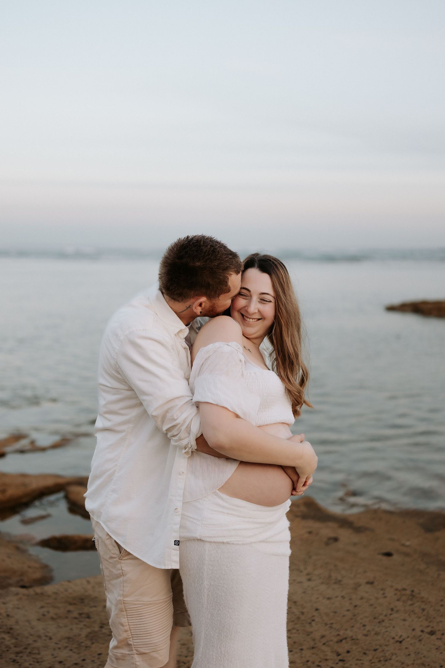 A couple embraces on a rocky beach at sunset, the man kissing the woman's cheek as she smiles warmly.