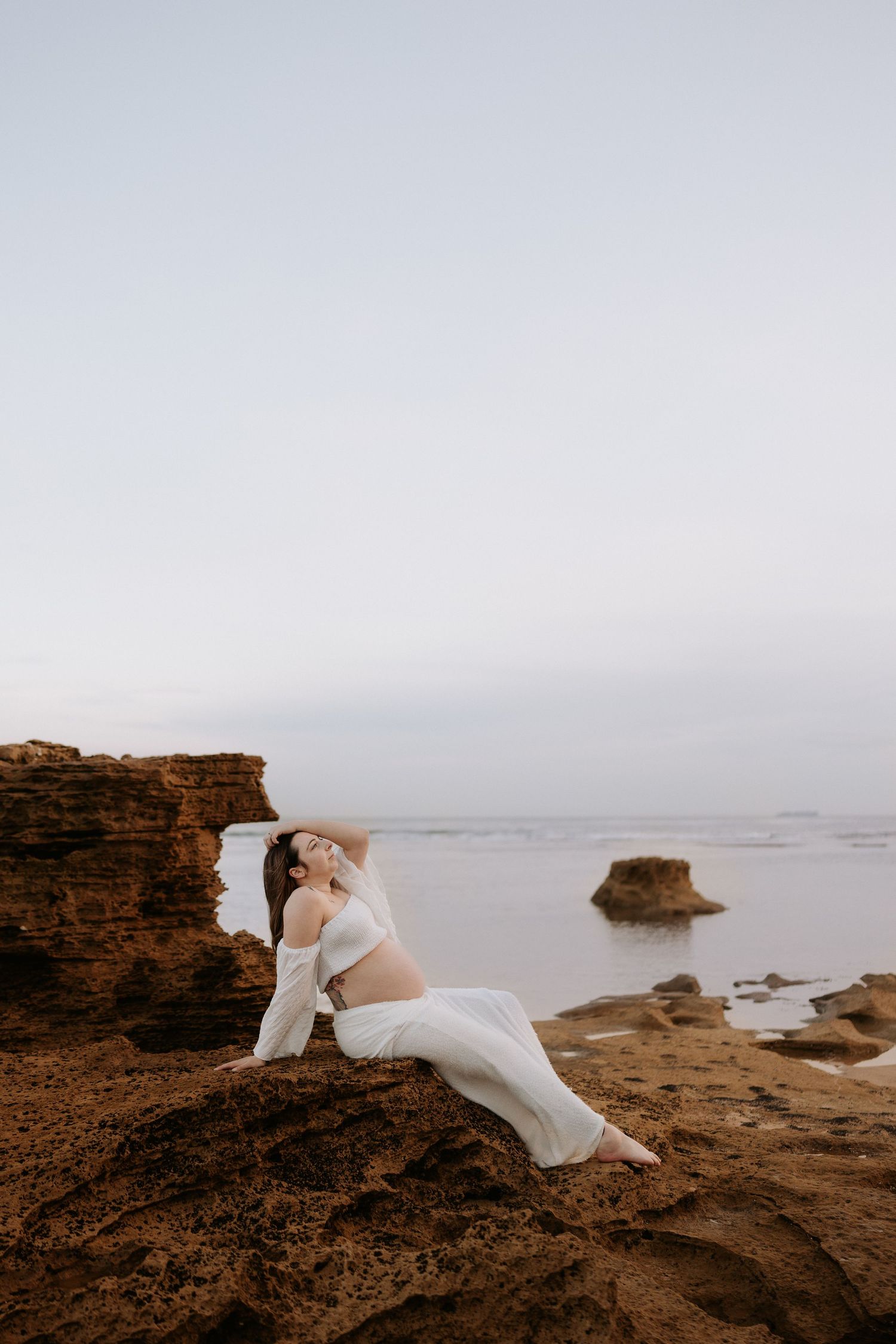 Pregnant woman in white dress poses gracefully on coastal rocks at dusk with ocean backdrop.