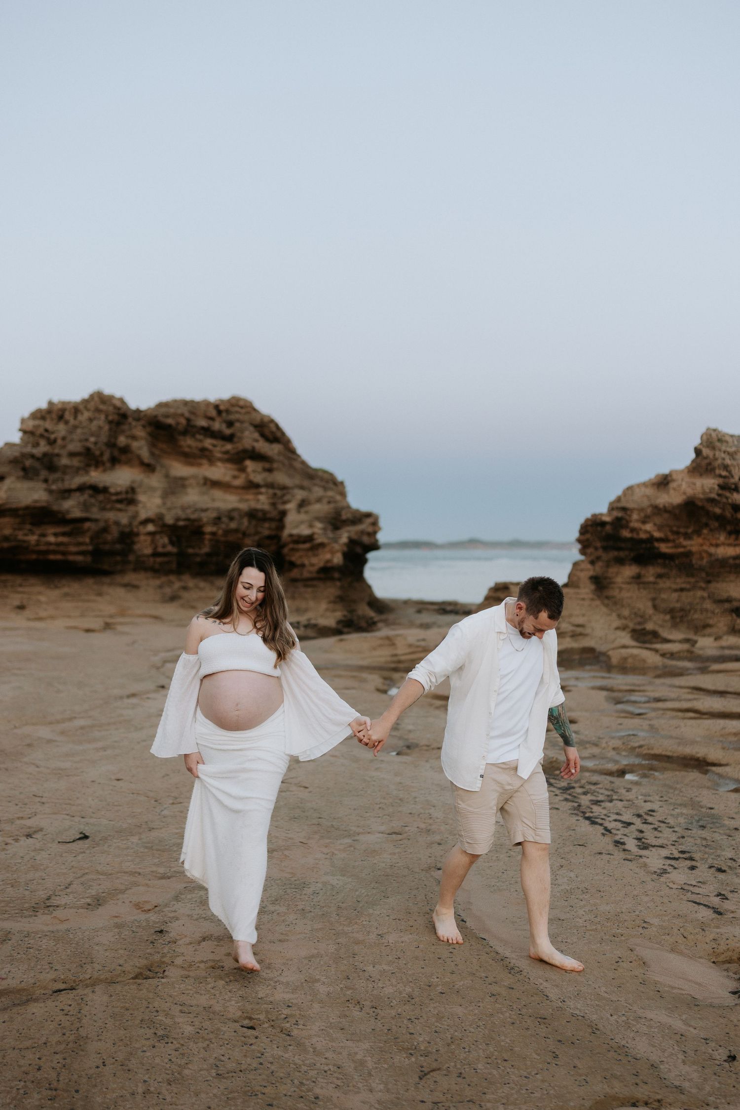Pregnant couple holding hands on a rocky beach, woman in white flowing dress showing baby bump, man in white shirt.