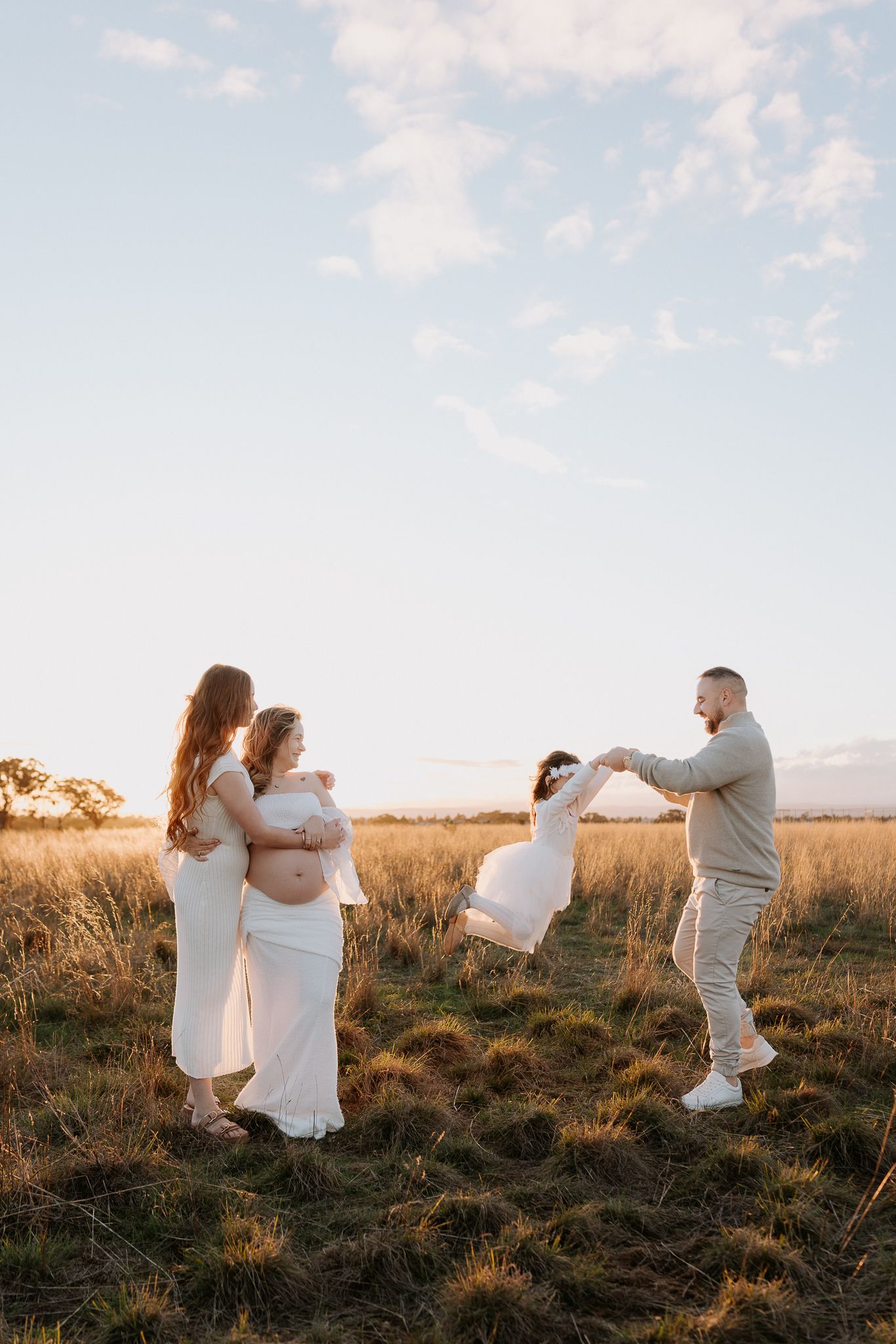 Pregnant woman in white dress with family at golden hour in grassy field, man swinging child while women embrace.