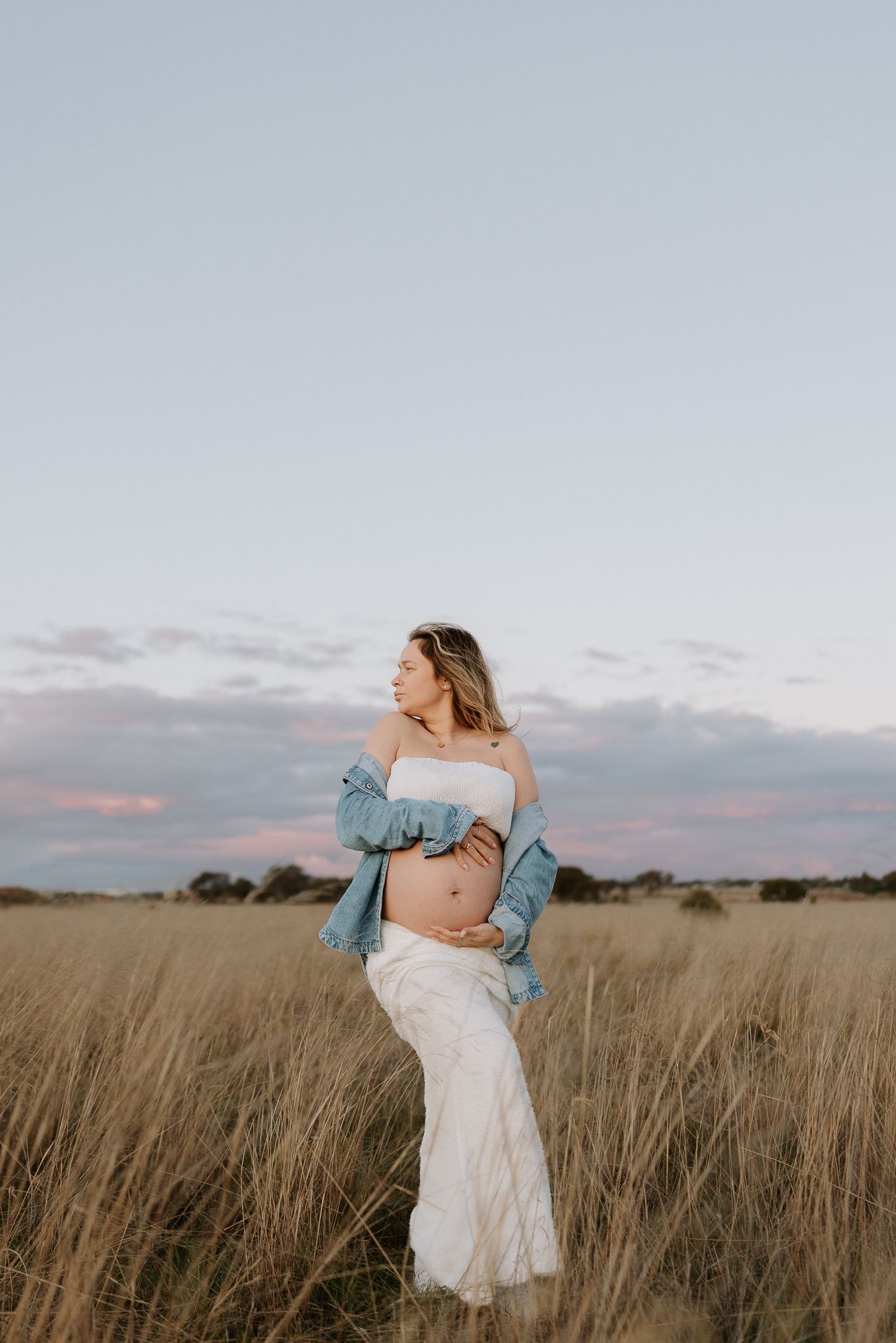 Pregnant woman in white dress and denim jacket cradling her baby bump in a golden grass field at sunset.