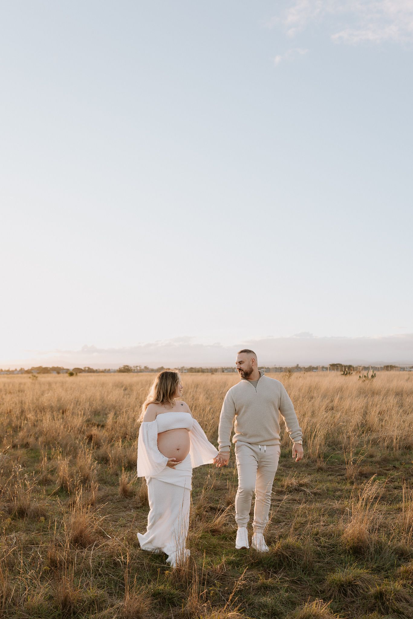 Pregnant couple holding hands in golden field at sunset, woman in white dress showing baby bump.