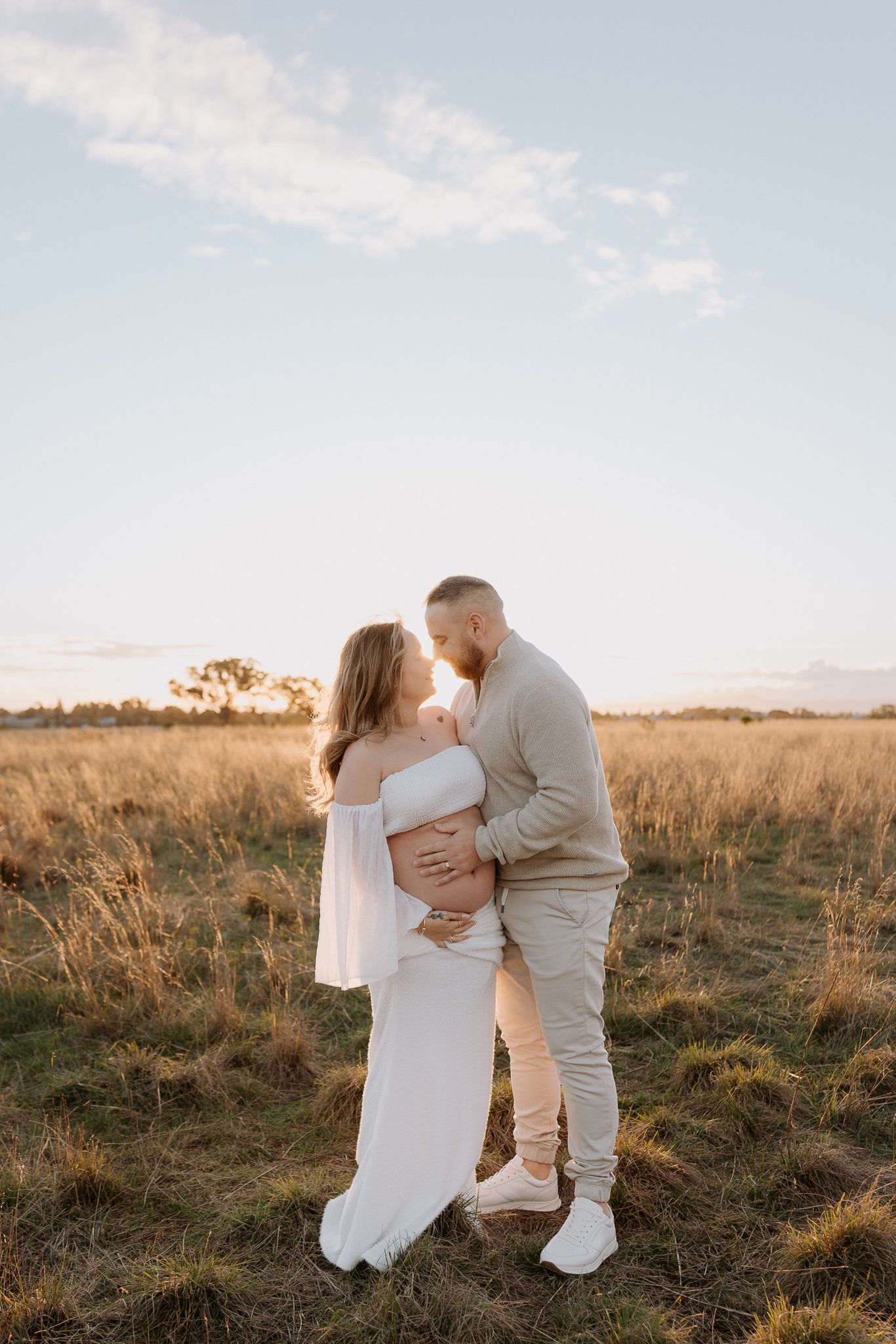 A couple shares a tender kiss in a golden field at sunset, the woman in a white dress cradling her pregnant belly.