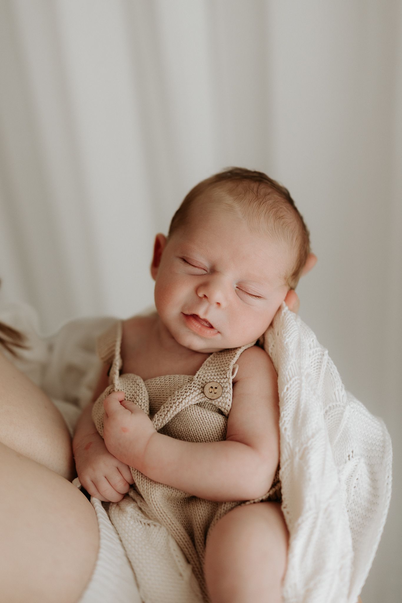 Newborn baby in beige knit romper resting in mother's arms with a white blanket.