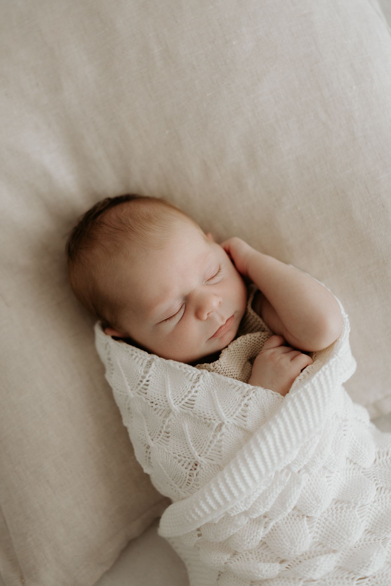 Peaceful newborn baby sleeping swaddled in a white knit blanket on a neutral linen background.