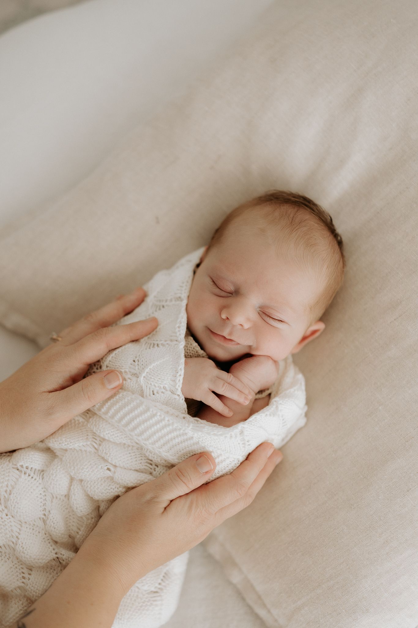 Sleeping newborn baby in white knit outfit cradled gently by mother's hands on a soft beige background.
