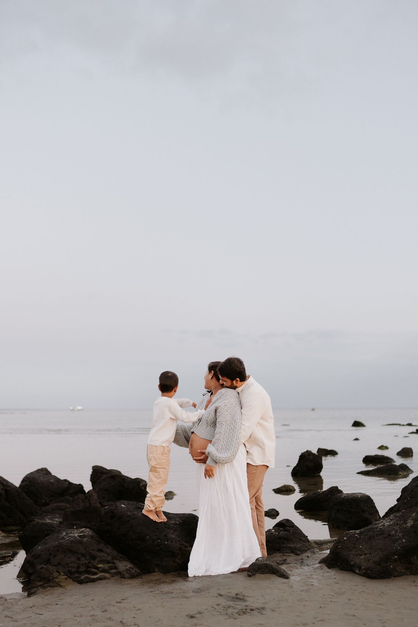 Family of three standing on rocky beach at dusk, parents embracing while holding young child, dressed in white and neutral tones.