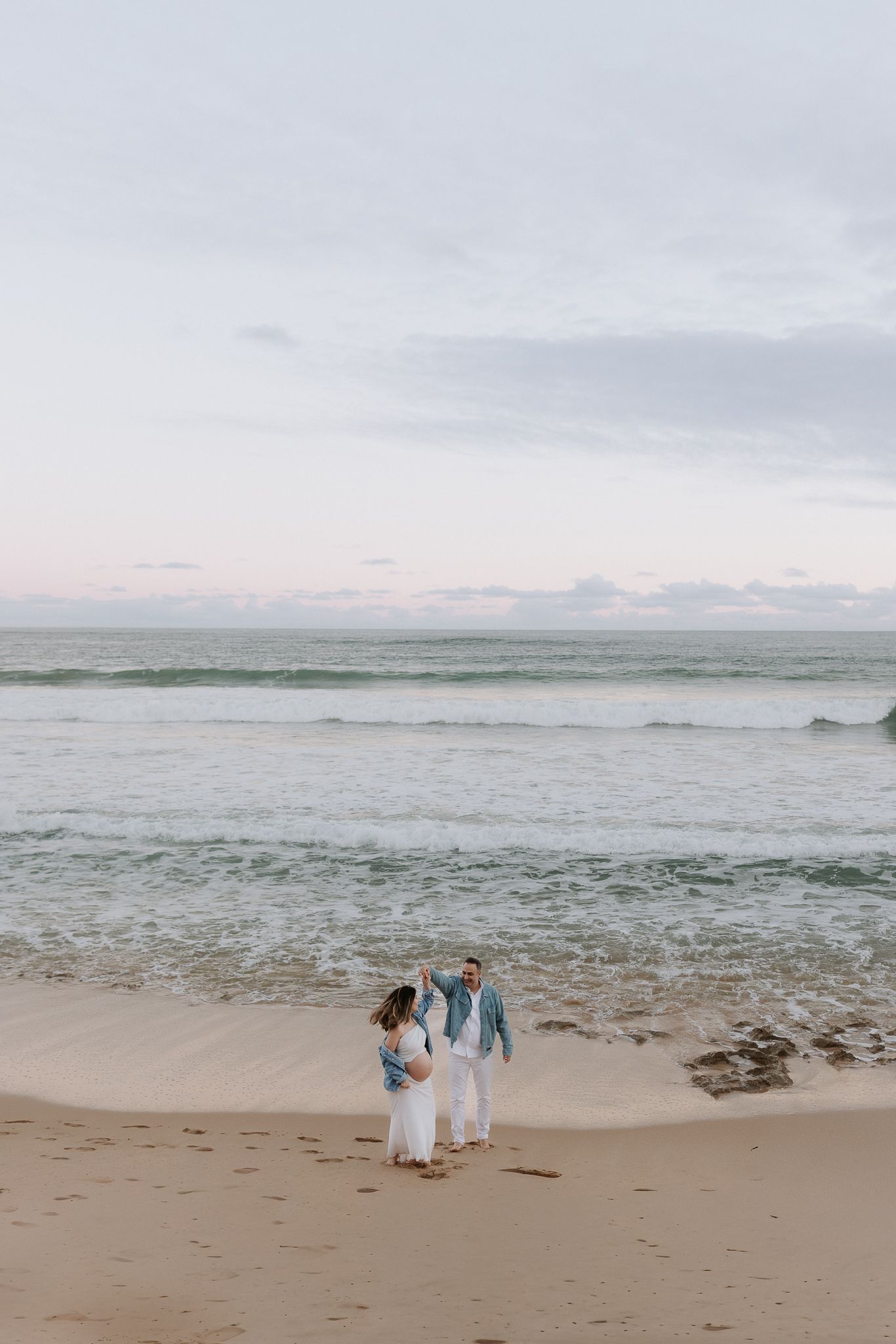 Couple dancing on a sandy beach at sunset with gentle ocean waves in the background.