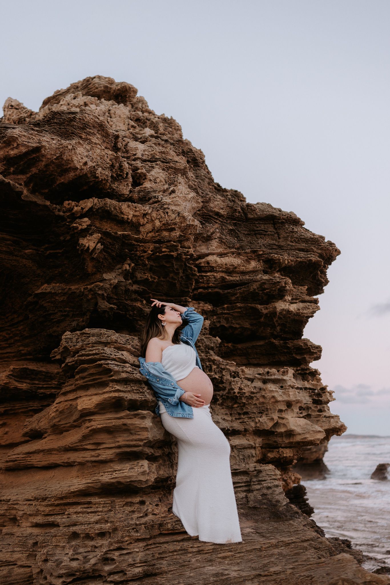 Pregnant woman in white dress and denim jacket poses against rugged coastal rock formation at dusk.