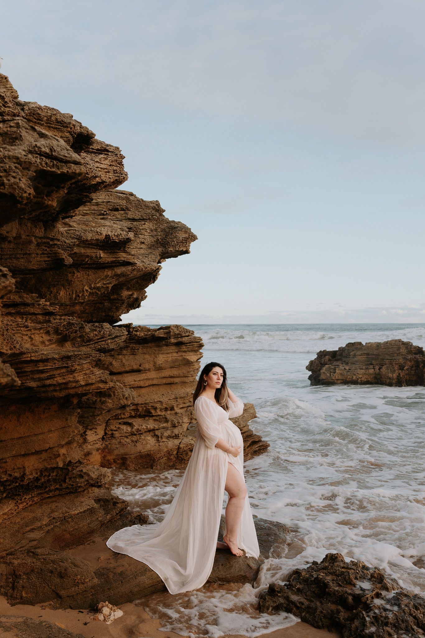 Pregnant woman in flowing white gown poses on rocky coastal cliffs with ocean waves crashing behind her.