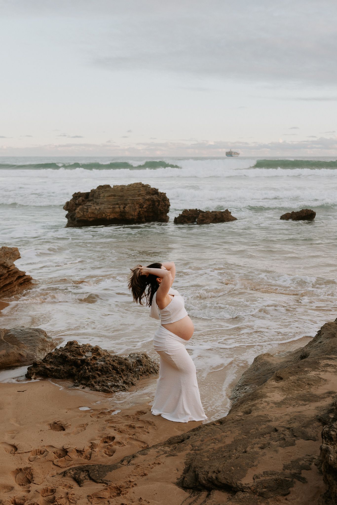 Pregnant woman in white outfit posing on sandy beach with rocky shoreline and ocean waves in background.