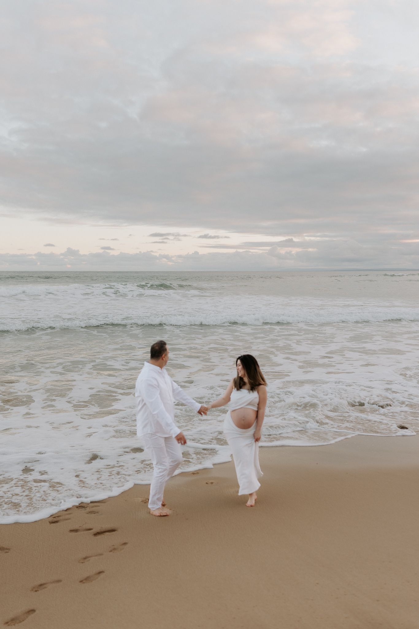 Pregnant couple in white outfits holding hands on a sandy beach with ocean waves at sunset.