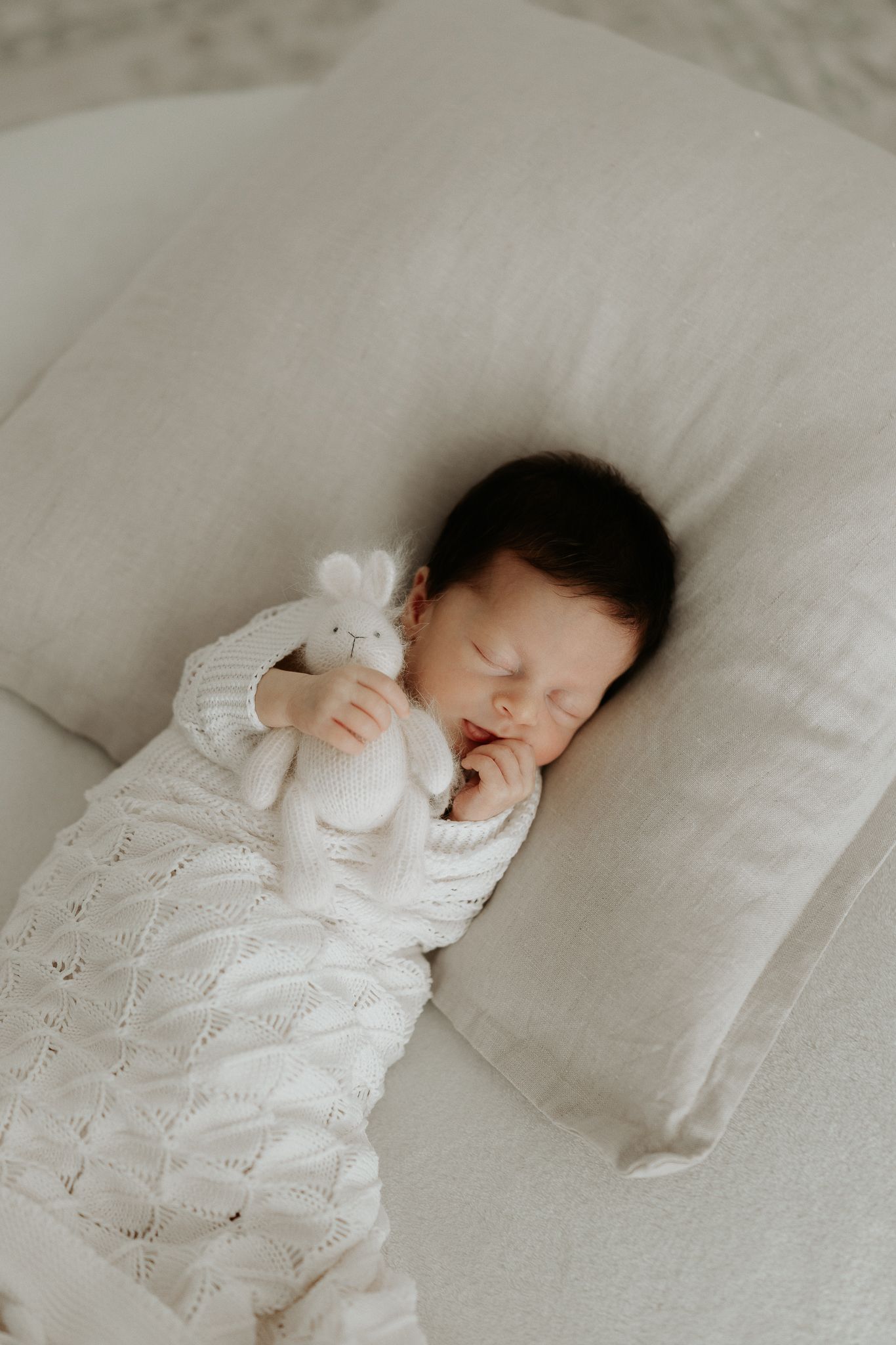 Sleeping newborn baby in white knit outfit holding a small bunny stuffed animal on a cream pillow.