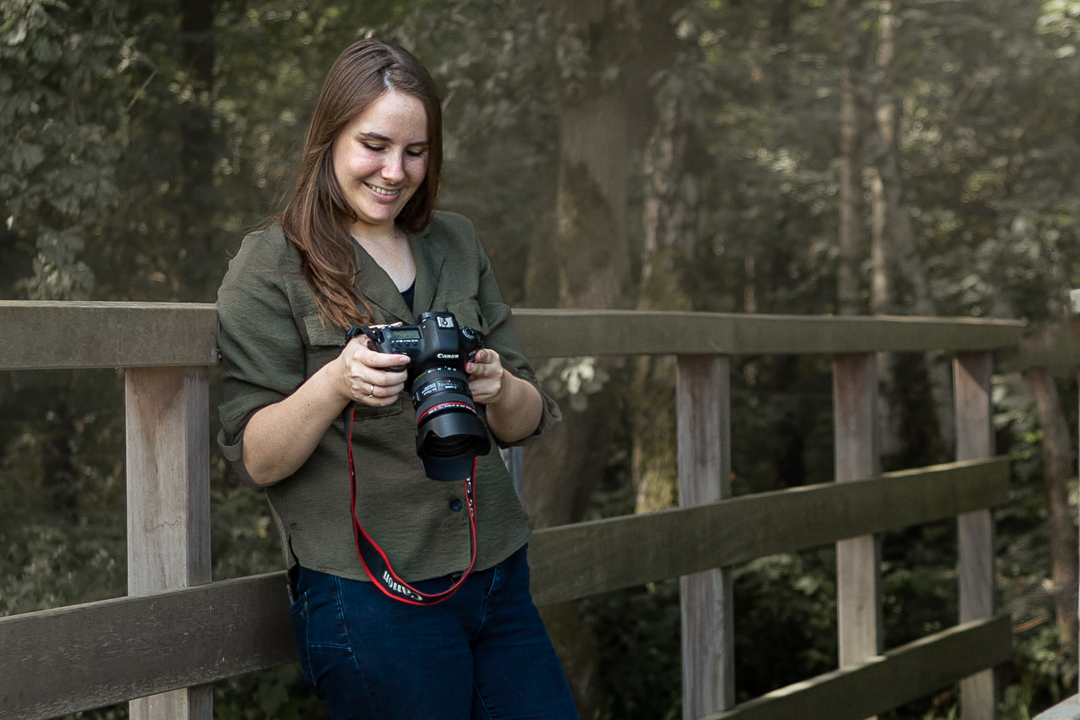 Fotografe staat buiten op een brug met camera in de handen, lachend omlaag kijkend.