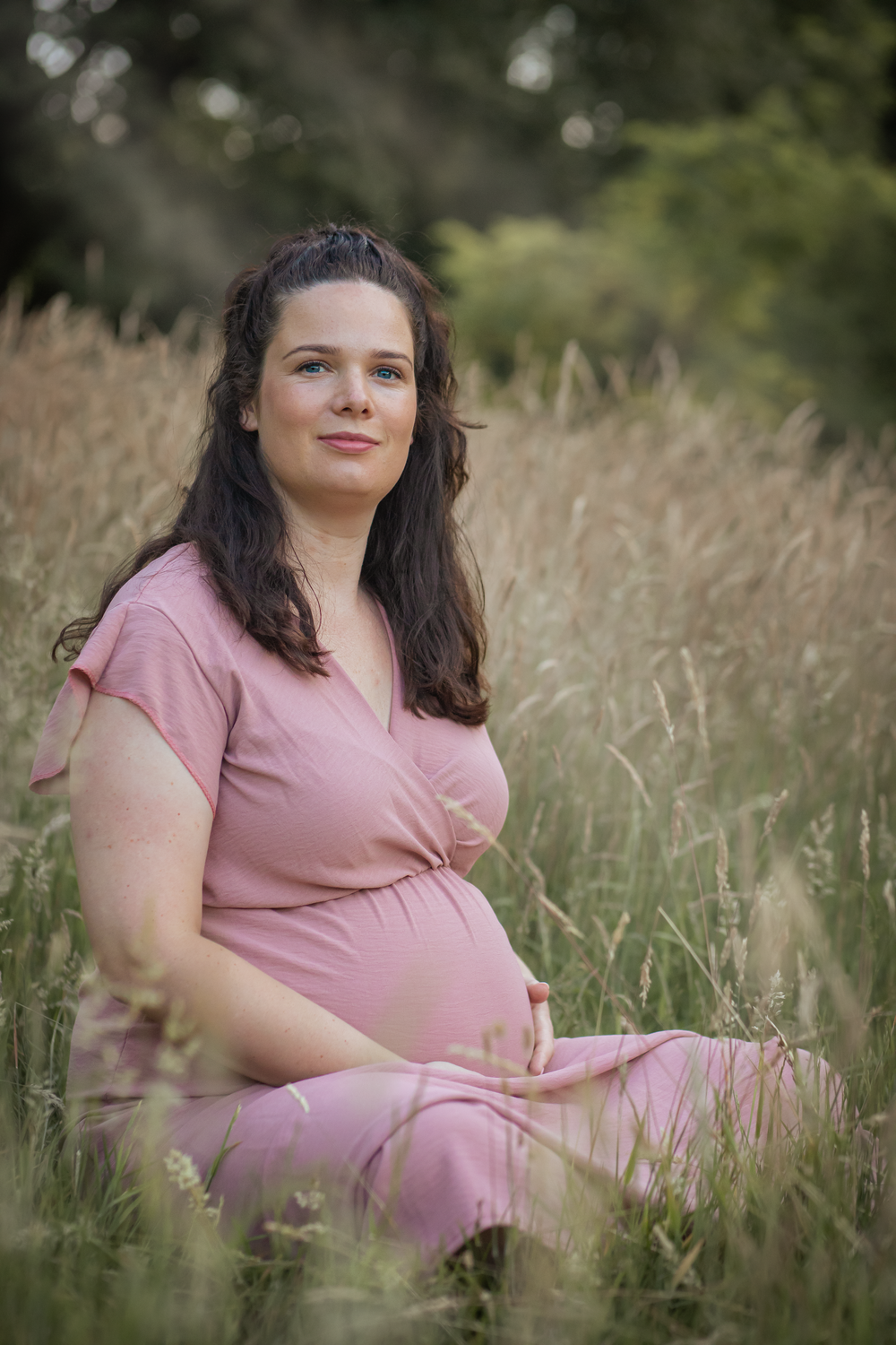 Zwangere vrouw in roze jurk zittend in een grasveld, handen op haar buik.