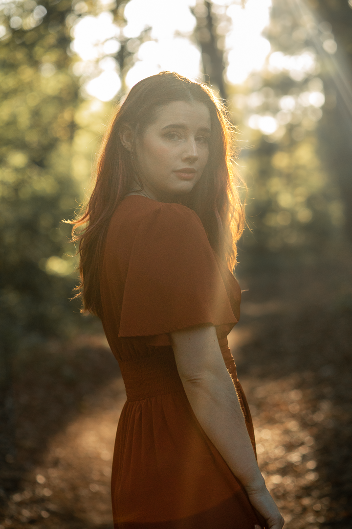 Vrouw in roest kleur jurk kijkt over haar schouder in een zonovergoten bos.
