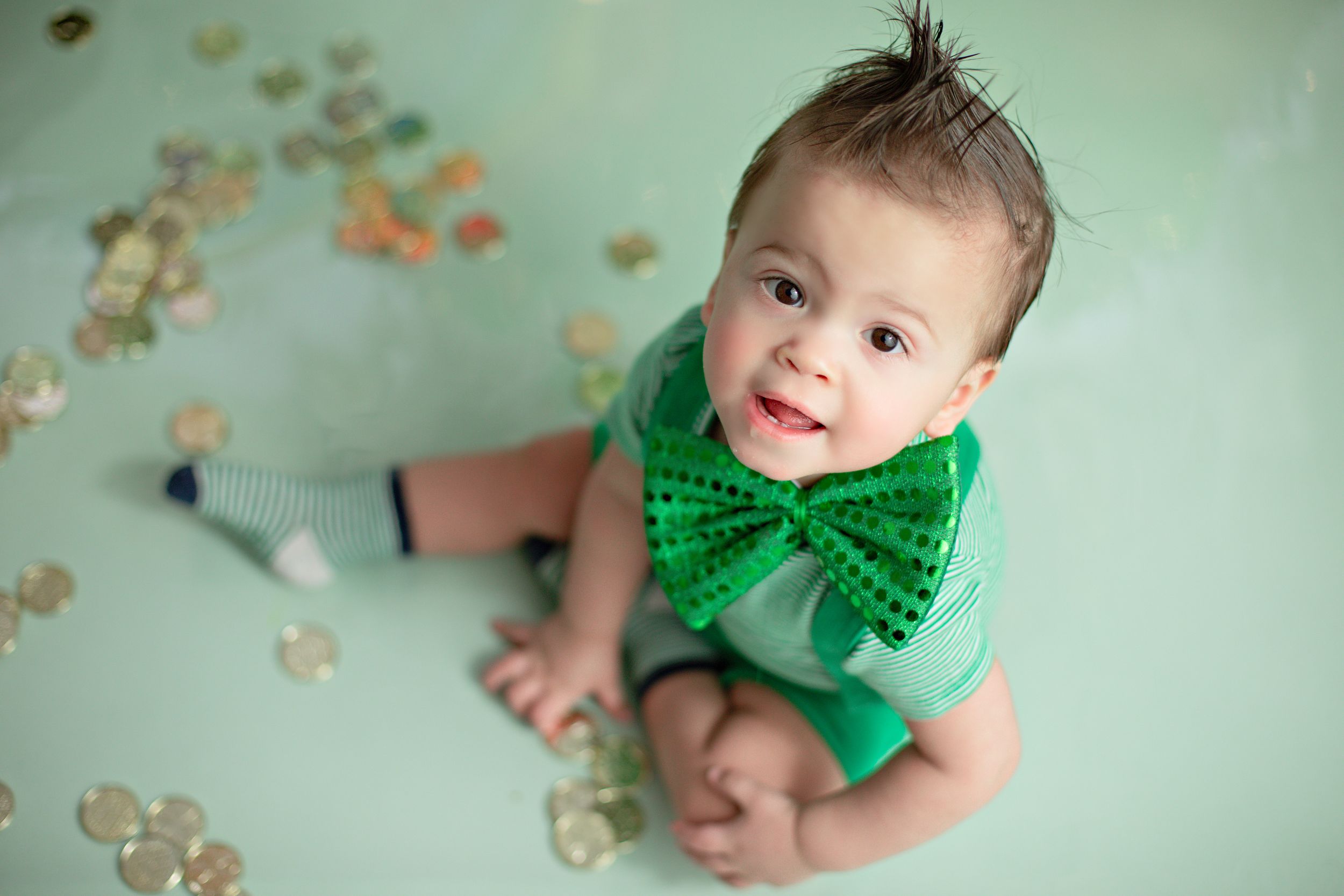 Baby in mint green outfit with sparkly green bowtie sits on floor with scattered confetti decorations around them.